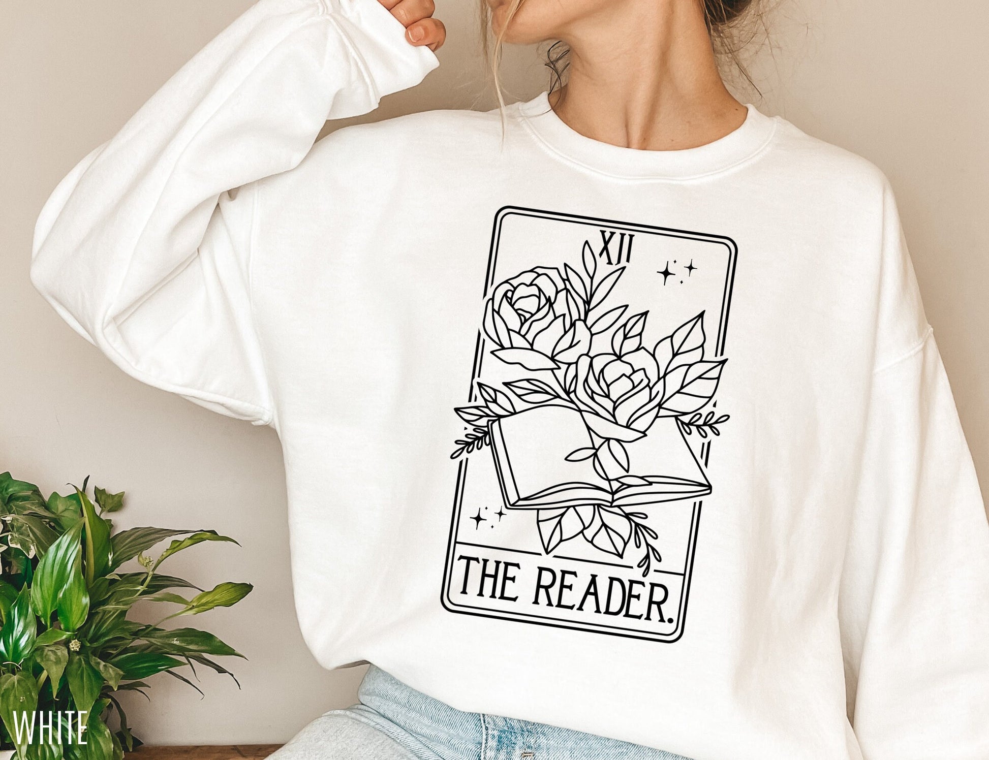 woman wearing white The Reader tarot sweatshirt with floral book graphic, sitting near plant
