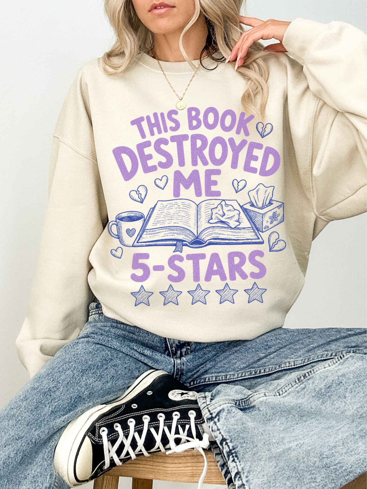woman wearing sand colored sweatshirt with this book destroyed me 5 stars graphic featuring open book coffee mug and heart illustrations for readers