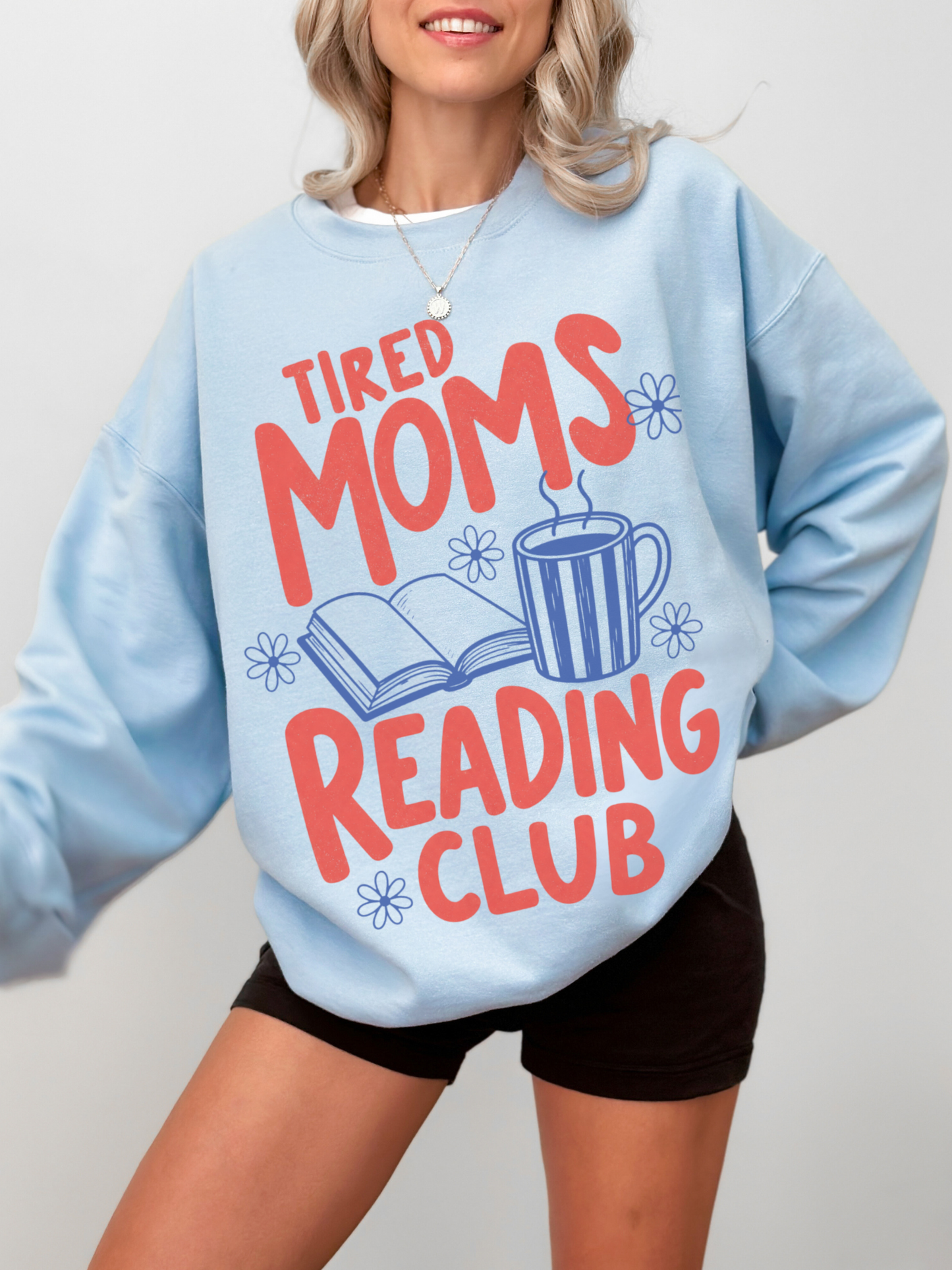 woman wearing oversized light blue tired moms reading club sweatshirt with retro typography coffee mug floral accents and open book artwork