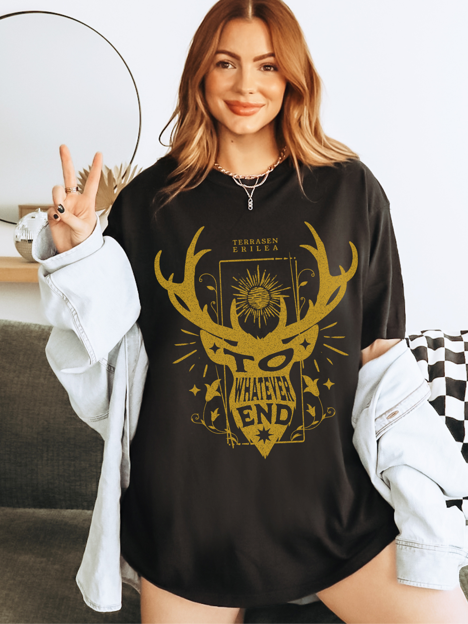 Woman wearing a black oversized “To Whatever End” shirt inspired by Terrasen and Erilea, styled with gold bookish fantasy design, posing with a peace sign in cozy home setting