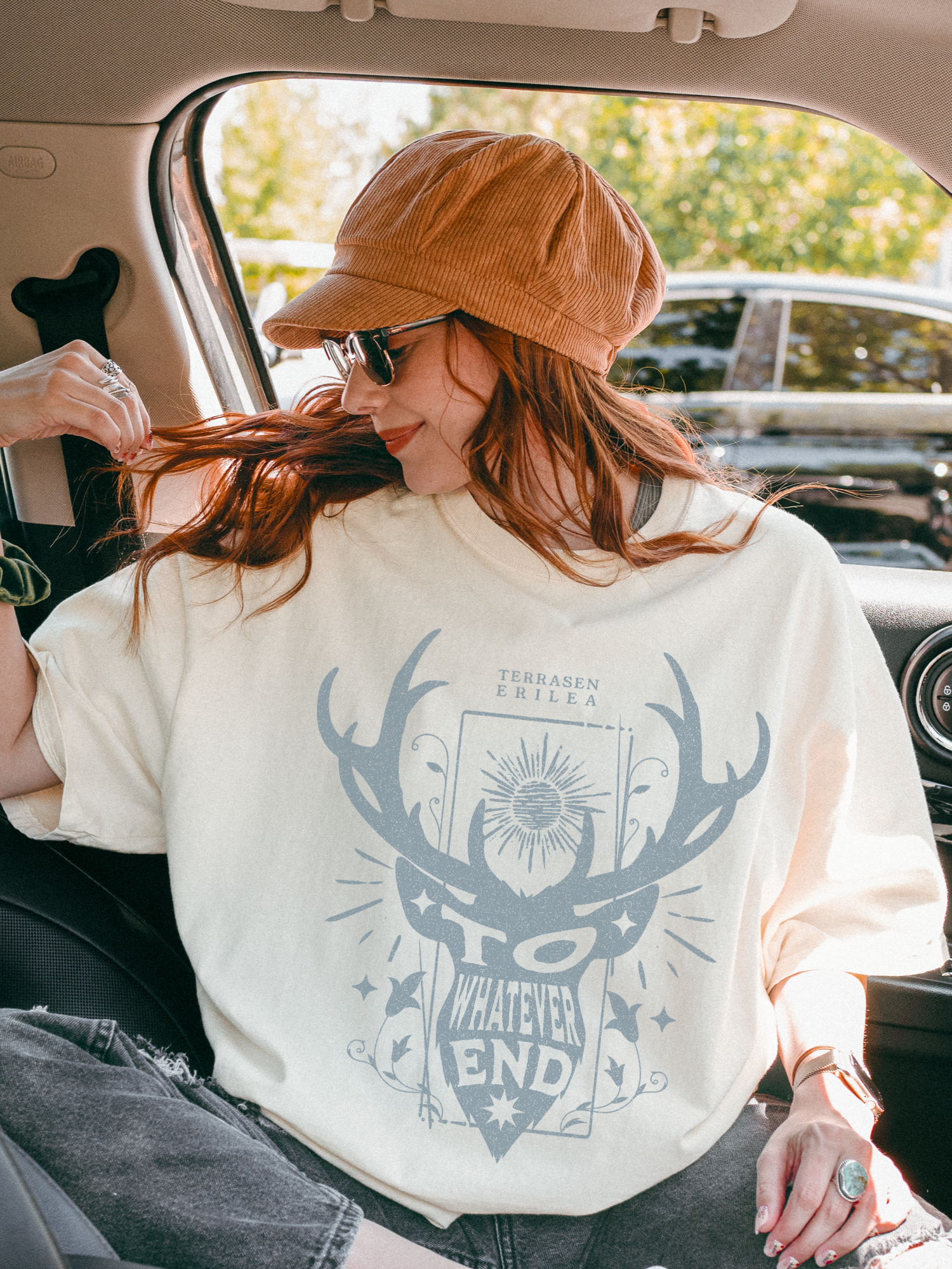 Red-haired woman in sunglasses and hat wearing a natural Terrasen shirt with a light blue “To Whatever End” design, sitting in a parked car
