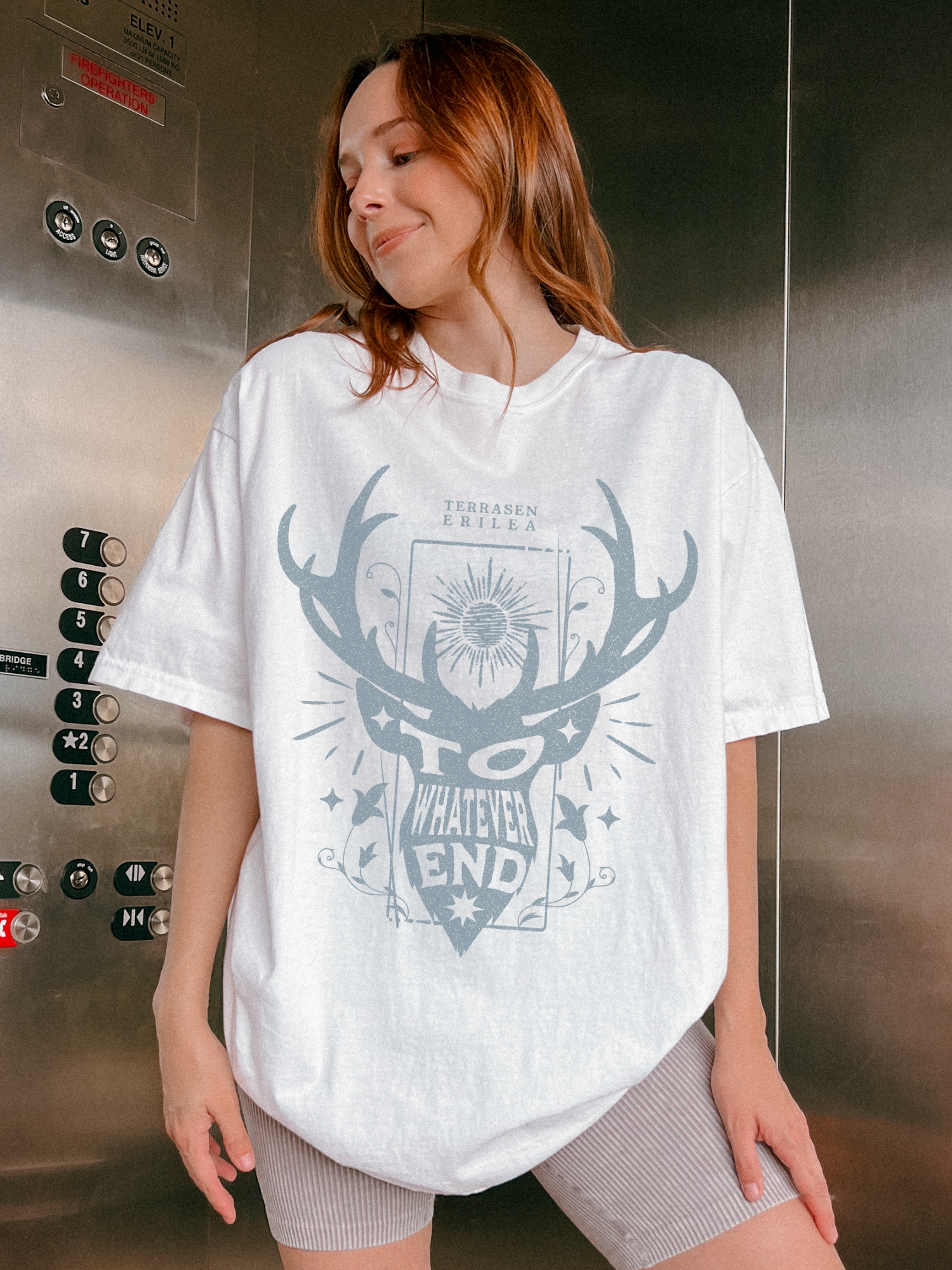 Red-haired woman in elevator wearing a white fantasy t-shirt with antler and sunburst artwork, inspired by Erilea