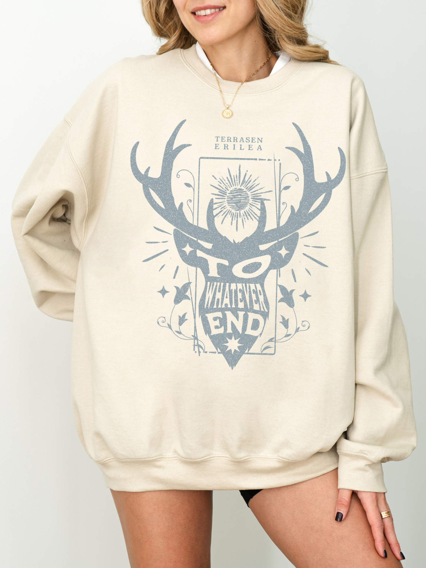 woman wearing sand-colored sweatshirt with blue antler crest and the quote “To Whatever End” from Throne of Glass
