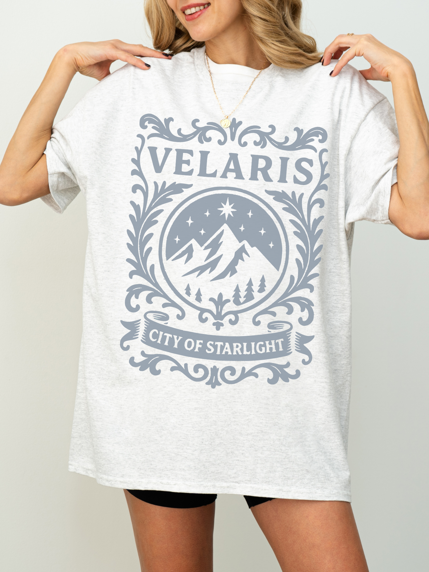 woman wearing an ash grey t-shirt with ornate design that says Velaris City of Starlight and features mountains and stars