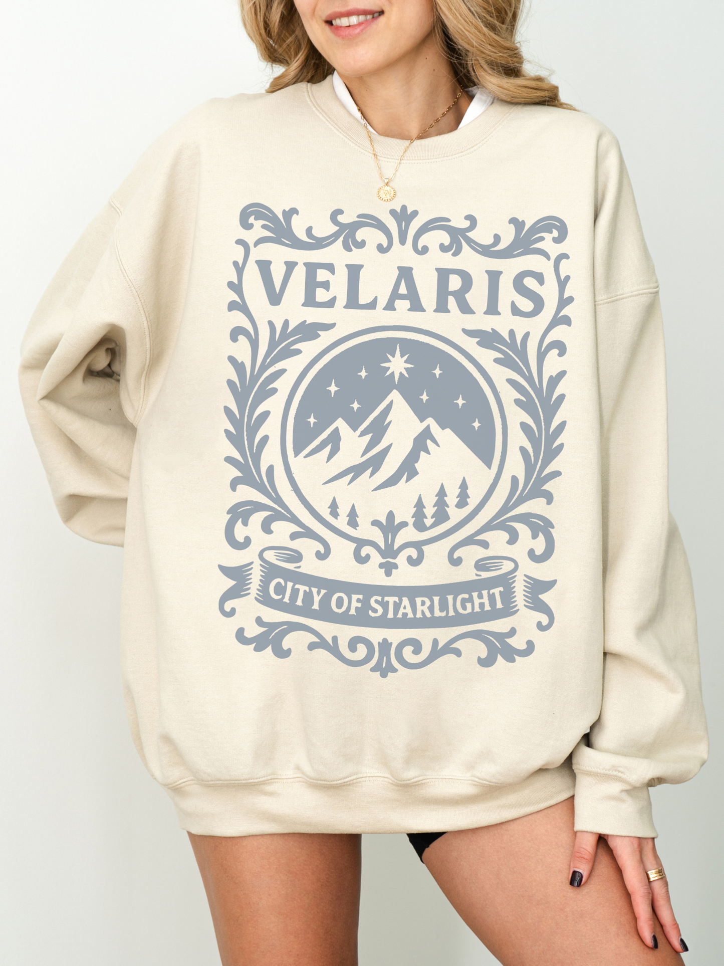 woman wearing a sand-colored sweatshirt with Velaris crest and City of Starlight text in blue grey ink