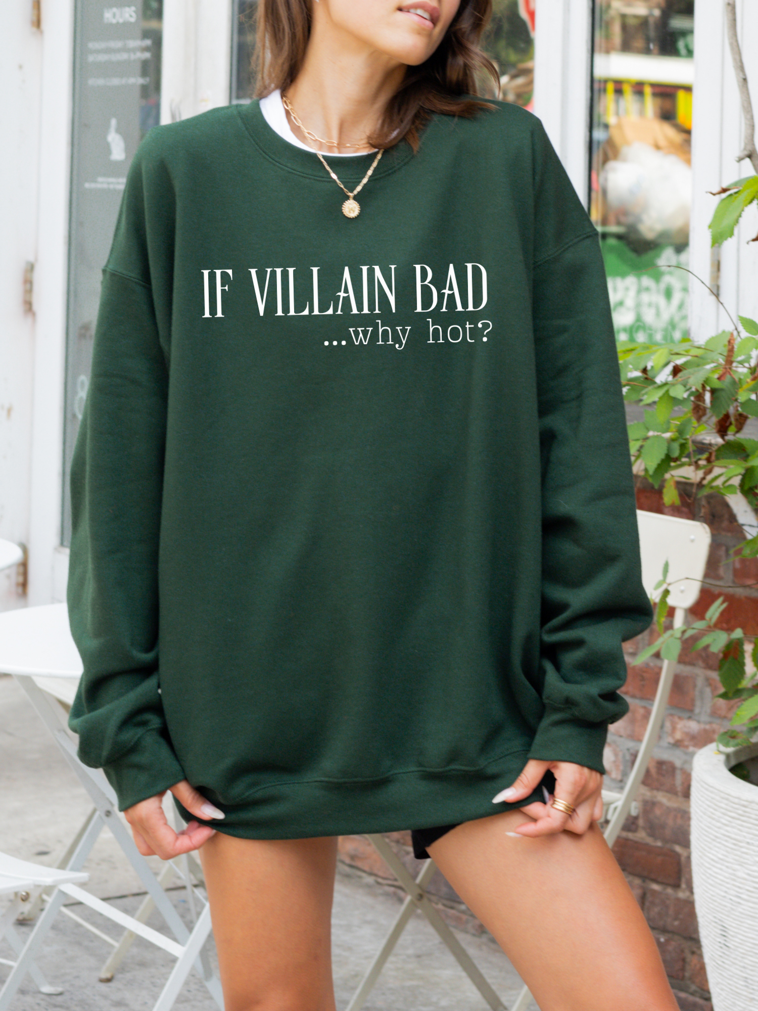 woman wearing a forest green sweatshirt that says if villain bad why hot standing outside near white chairs