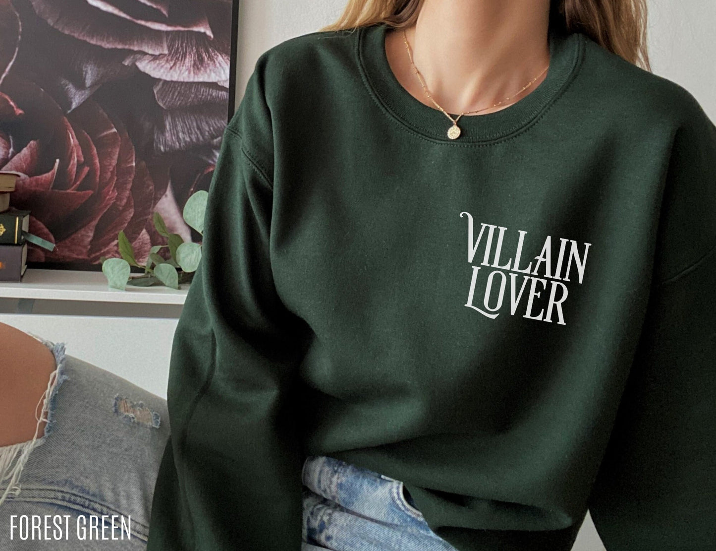 woman wearing forest green sweatshirt that says villain lover, seated by dark floral artwork and plants