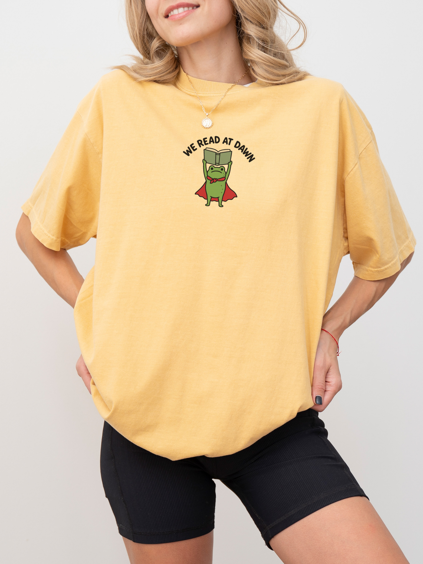 woman wearing a mustard Comfort Colors shirt featuring a reading frog superhero and We Read At Dawn design for avid readers