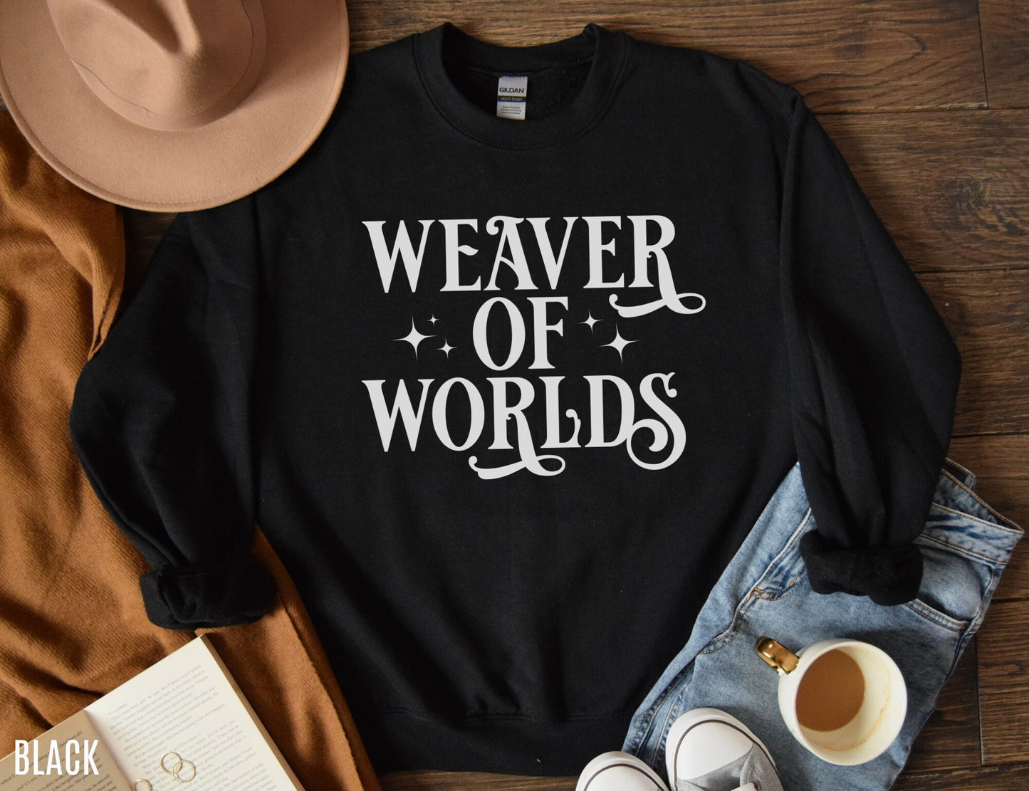 folded black sweatshirt with white “Weaver of Worlds” print, laid out with jeans, hat, coffee, and a book