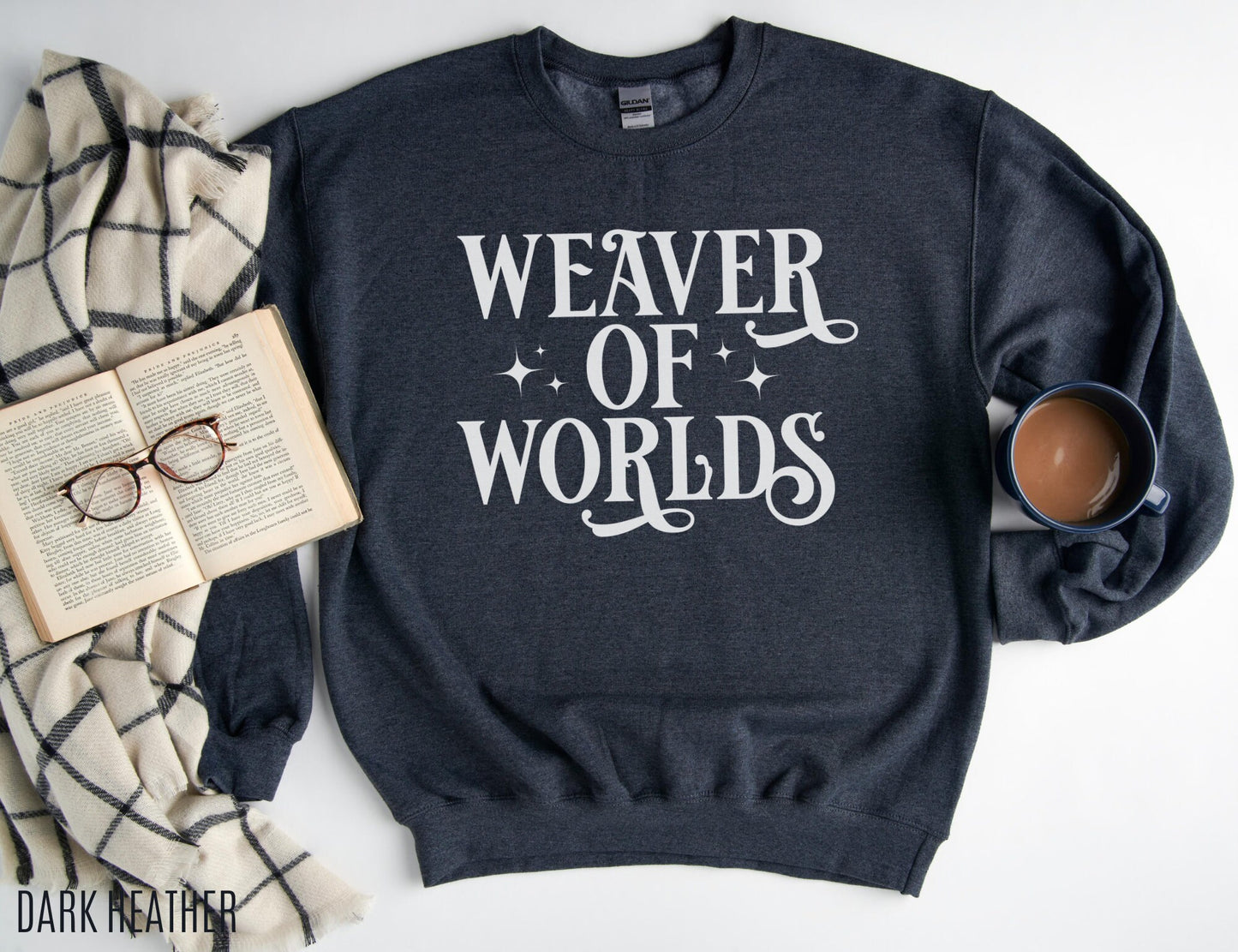 dark heather grey sweatshirt laid flat with white “Weaver of Worlds” text, open book, glasses, and a coffee mug
