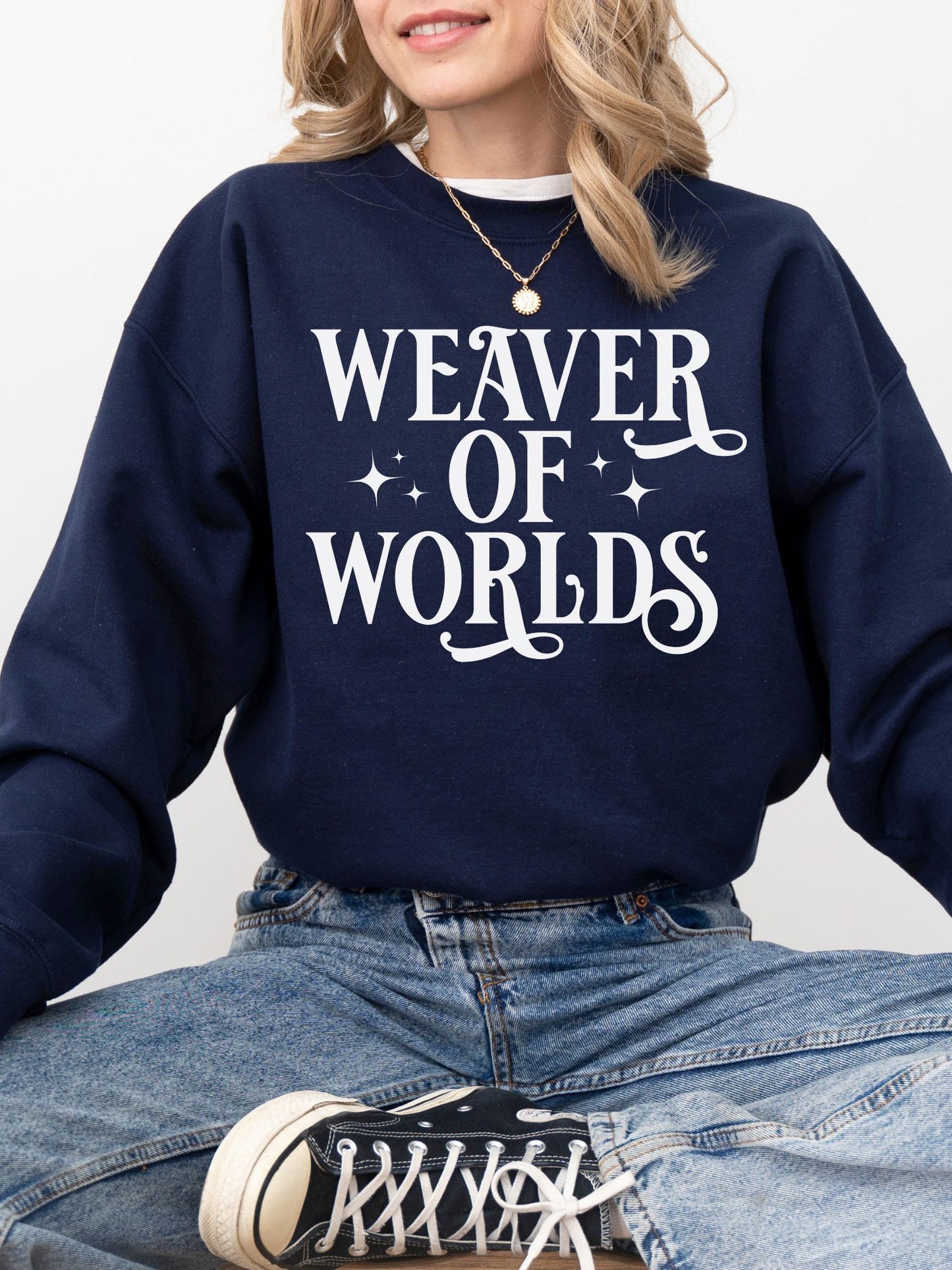woman in navy blue sweatshirt with whimsical “Weaver of Worlds” design, sitting cross-legged in jeans