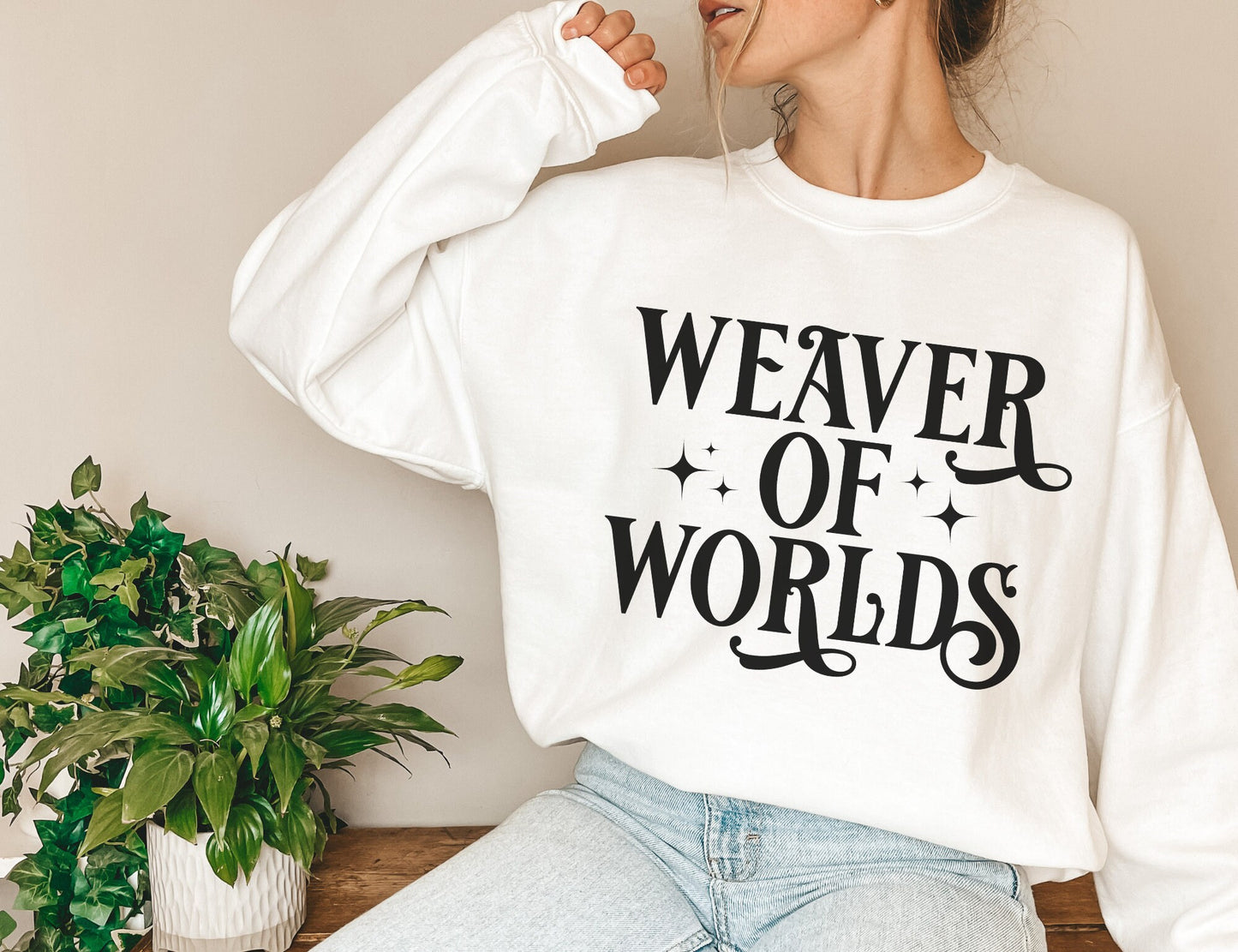 woman in white sweatshirt with fantasy-inspired “Weaver of Worlds” text and stars, sitting near green plants