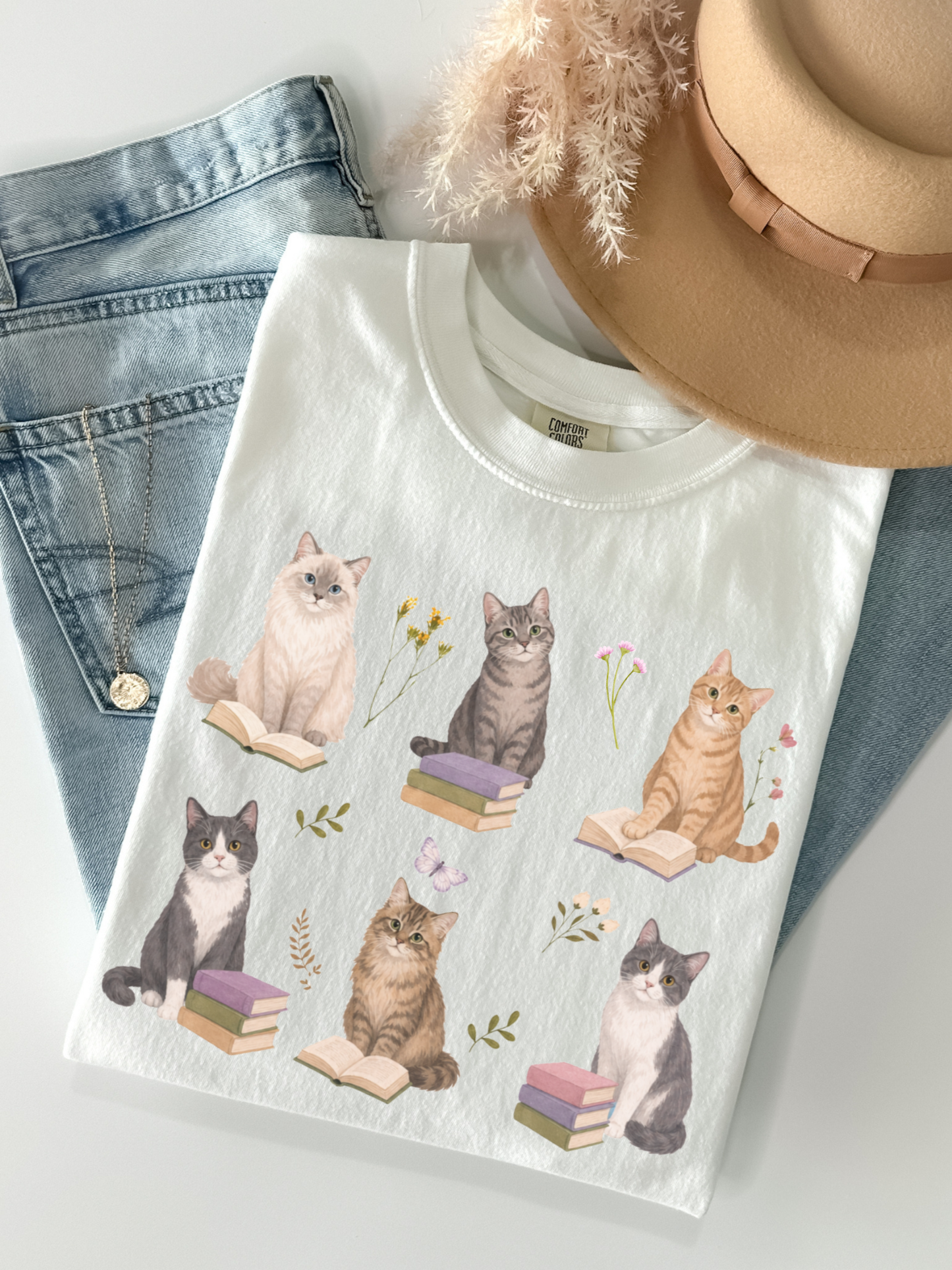 Flat lay of white Comfort Colors t shirt featuring cats reading books with floral accents styled with denim jeans gold necklace and beige hat