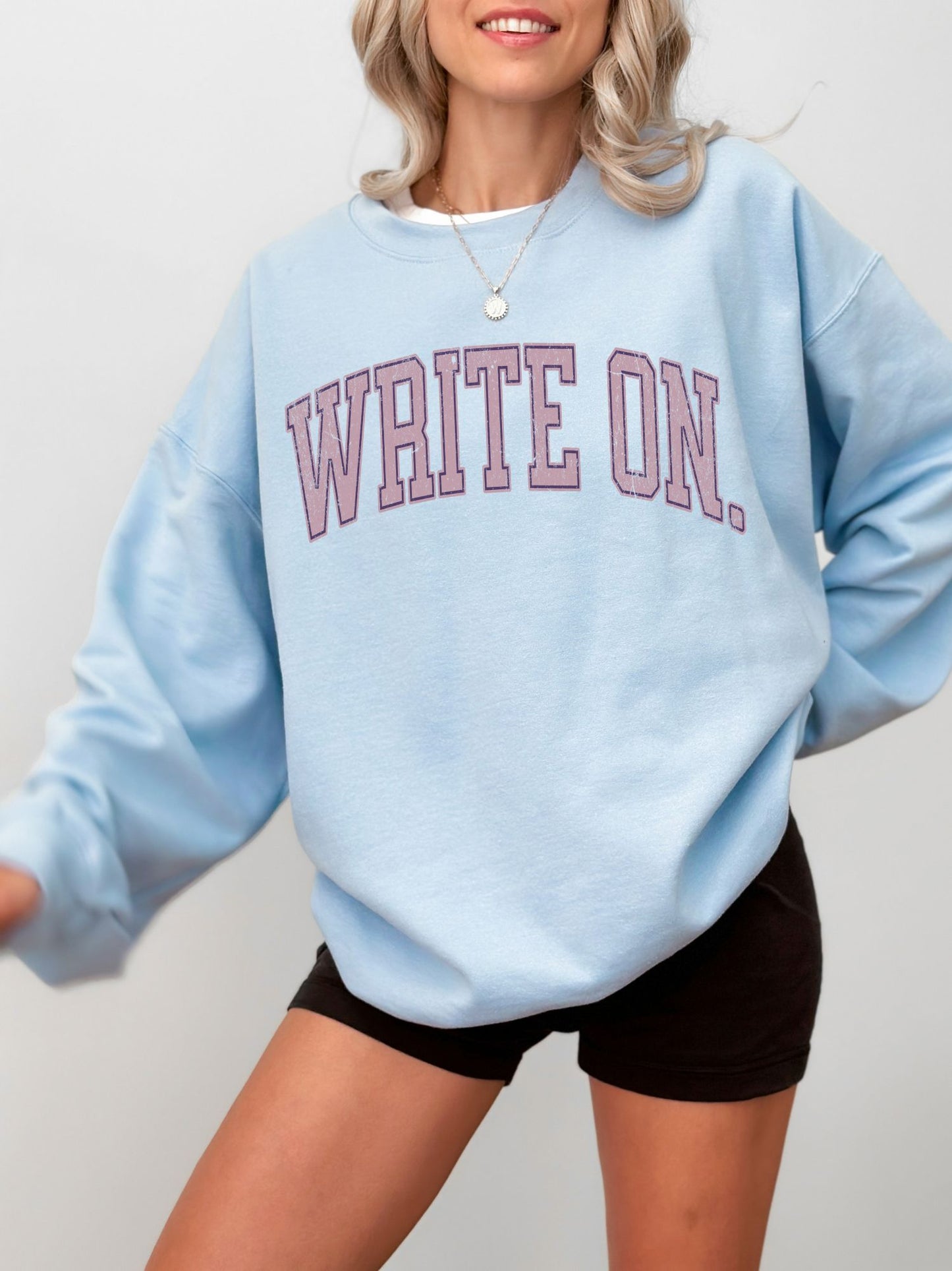woman wearing light blue sweatshirt with dusty rose write on text and black shorts