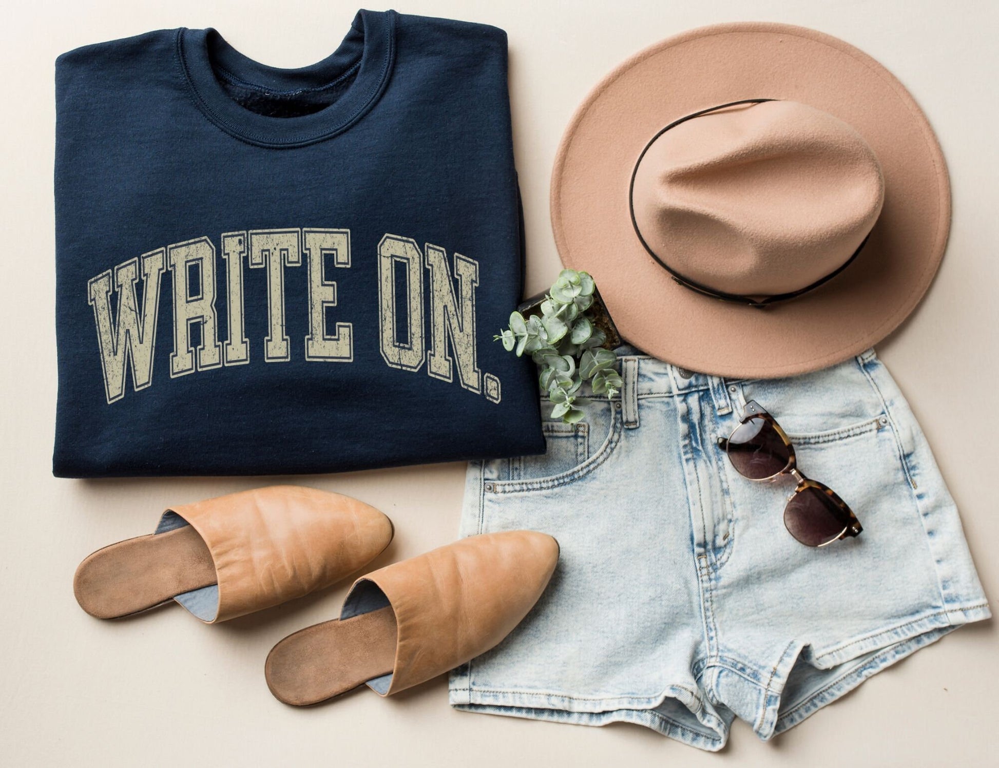 flatlay of folded navy write on sweatshirt with distressed cream text, paired with shorts, hat, and sunglasses