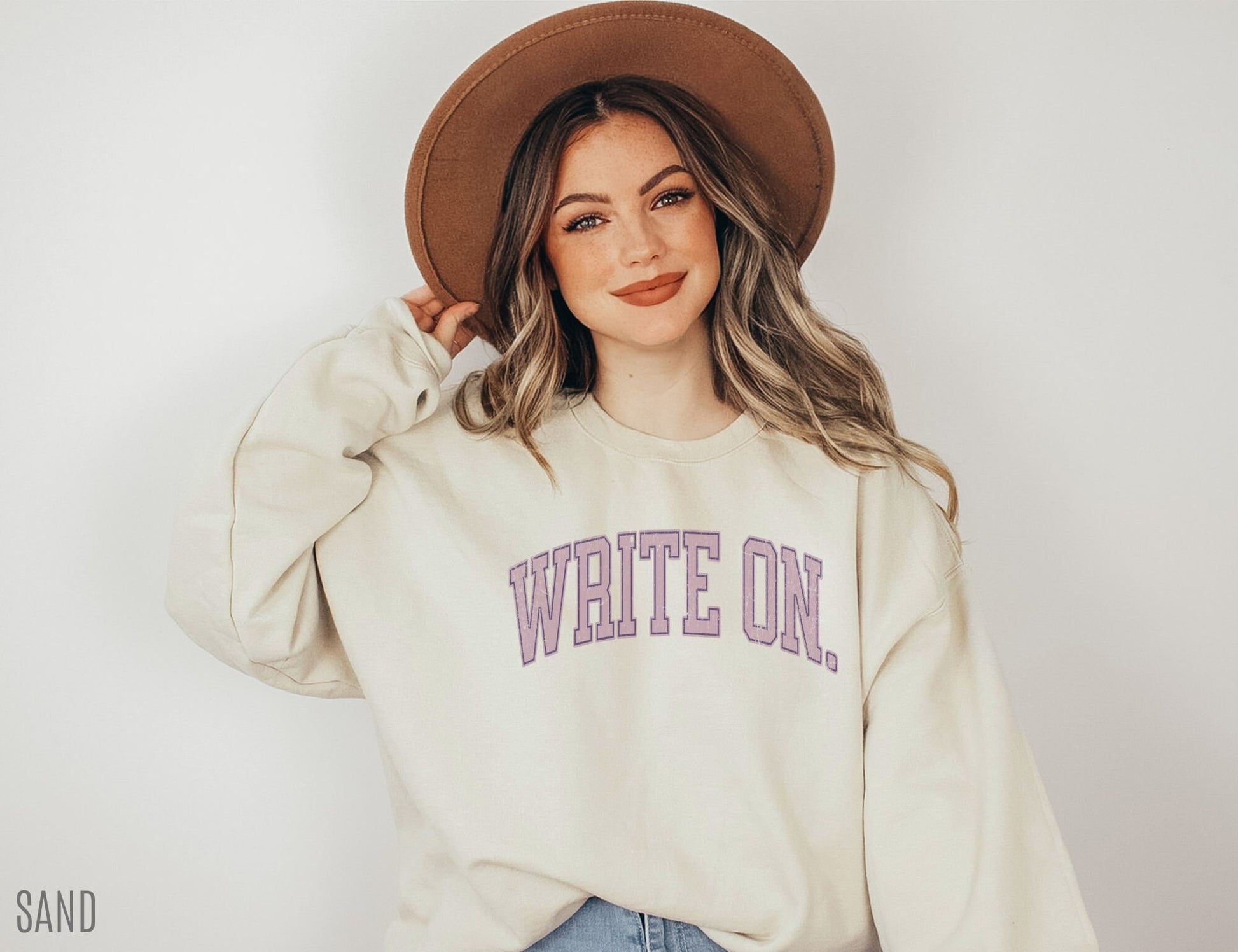 smiling woman in sand-colored sweatshirt with lavender write on text and wide-brim hat