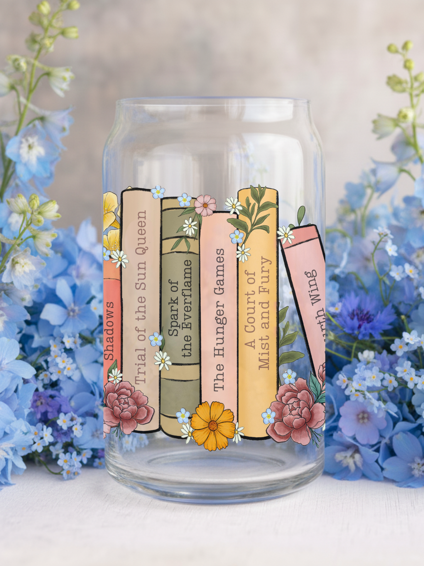 Clear glass tumbler featuring pastel-colored book spines like “A Court of Mist and Fury” and “The Hunger Games,” surrounded by hand-drawn florals and blue blossoms for a dreamy book lover aesthetic.