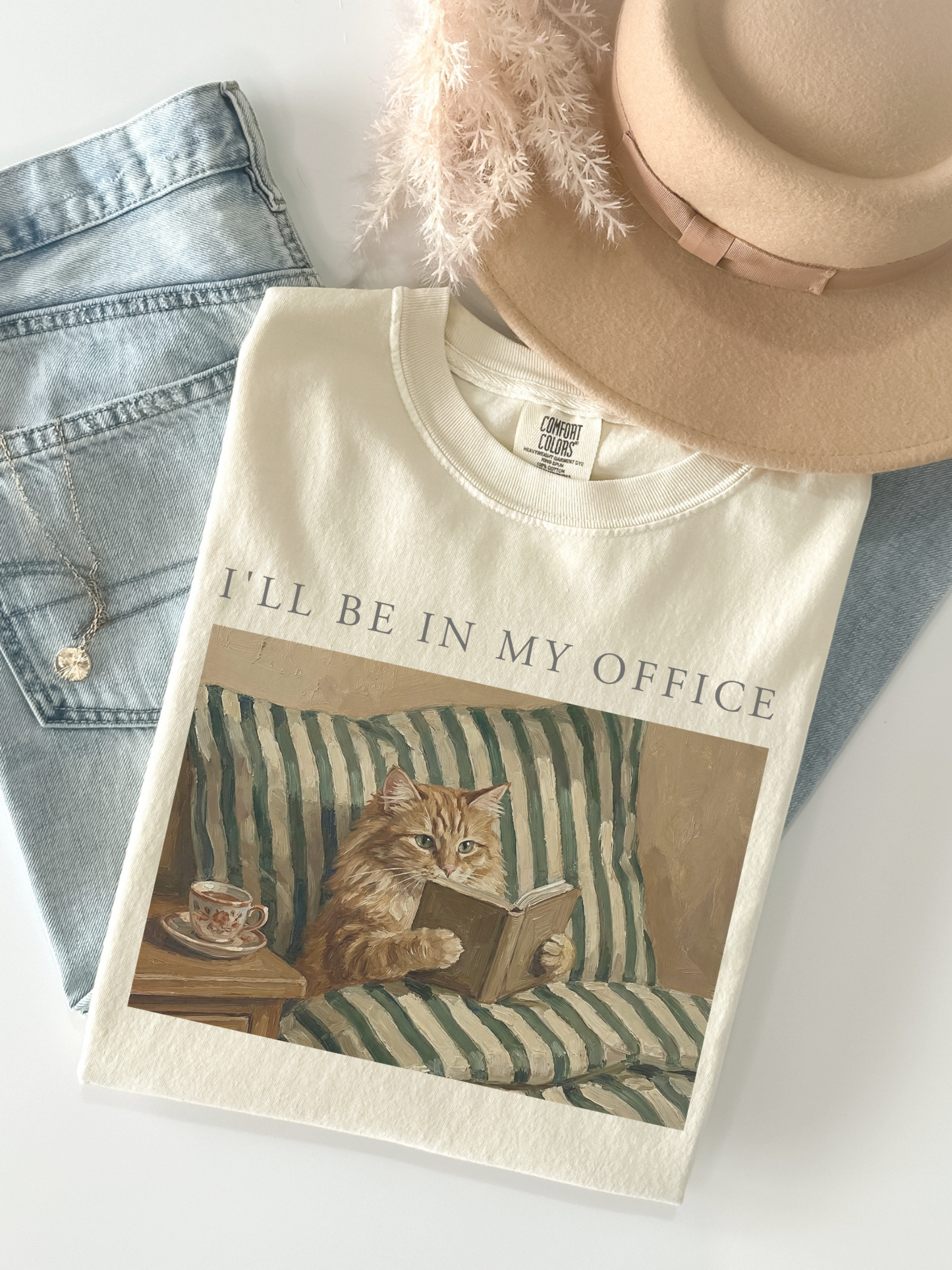 Flat lay of a ivory comfort colors t shirt with Ill be in my office text and a vintage painting of a cat reading a book in bed beside a tea cup. Styled with denim jeans and a neutral hat creating a cozy book lover aesthetic product photo.