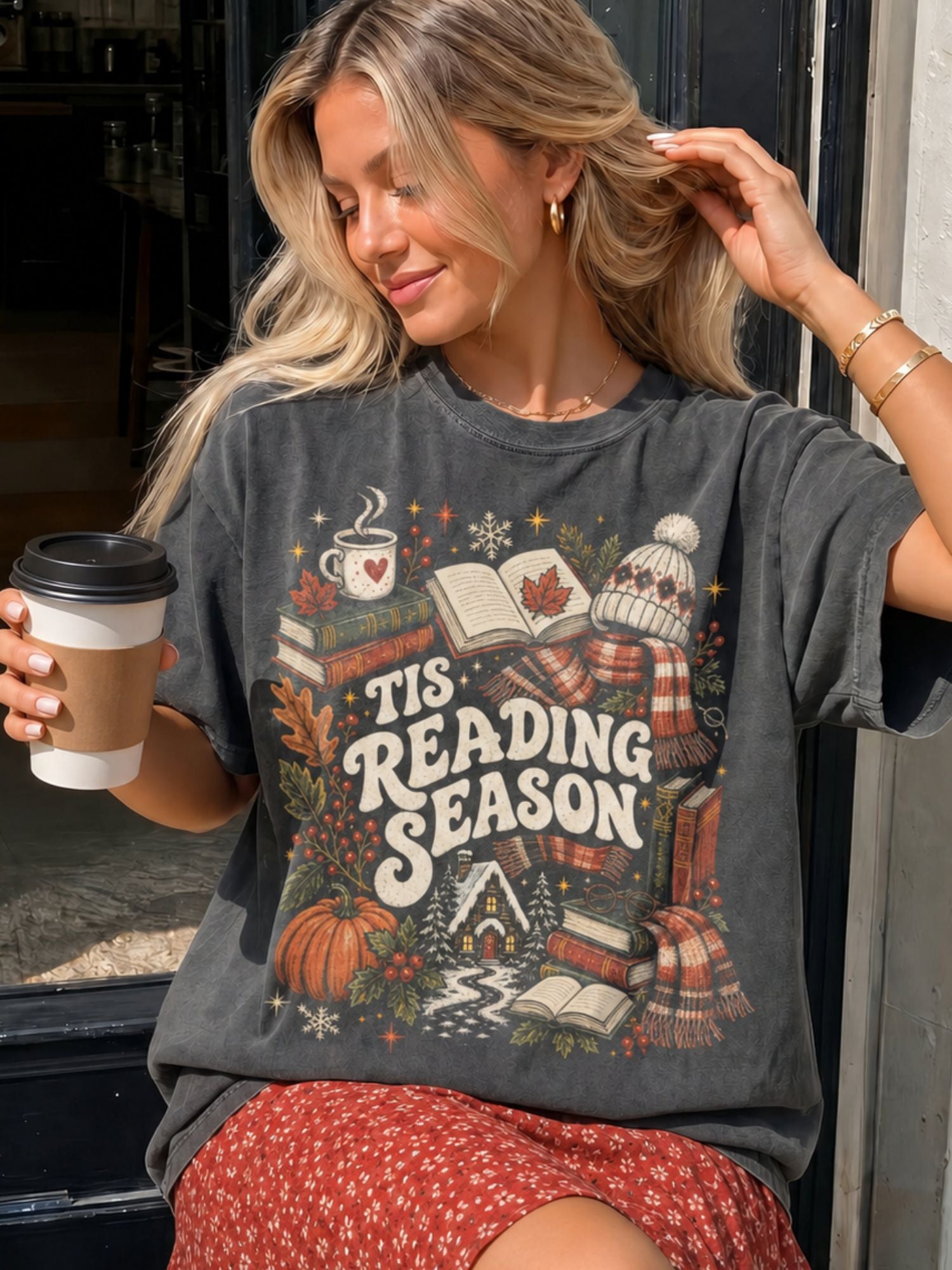 woman sitting outside a coffee shop, wearing a pepper tshirt that says "tis reading season" with christmas and reading icons around it