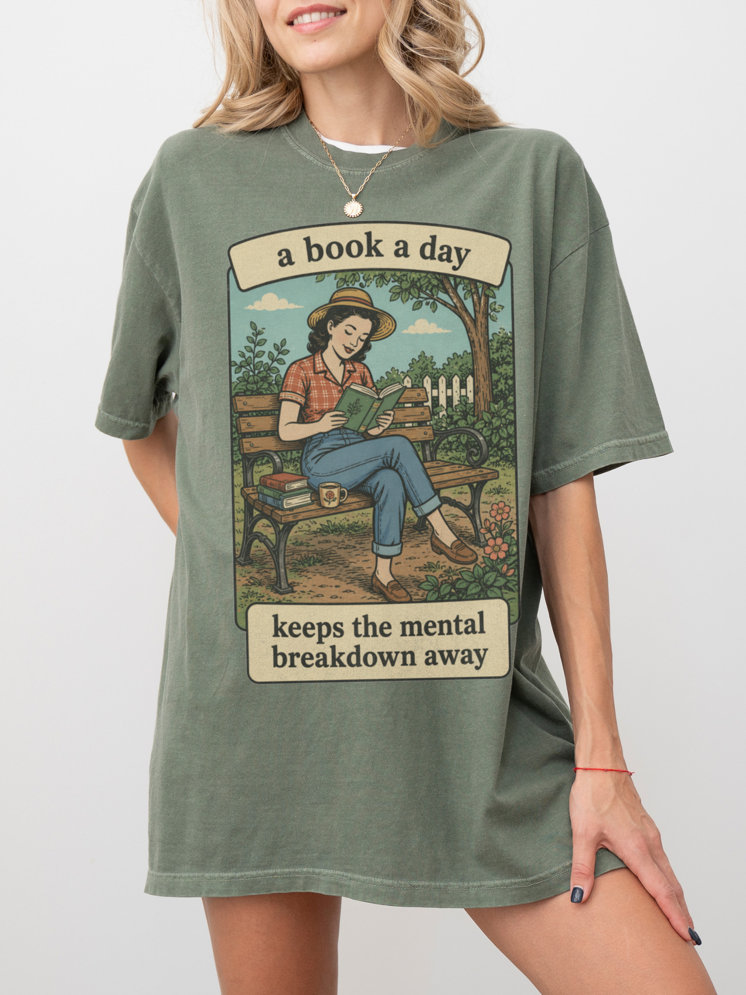 Woman wearing a moss green oversized t shirt with vintage style illustration of woman reading outdoors and text a book a day keeps the mental breakdown away