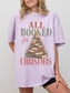 Orchid Comfort Colors shirt featuring an All Booked for Christmas design with a vintage book Christmas tree made from stacked red and green books, evergreen branches, ornaments, berries and gold star, festive reader shirt