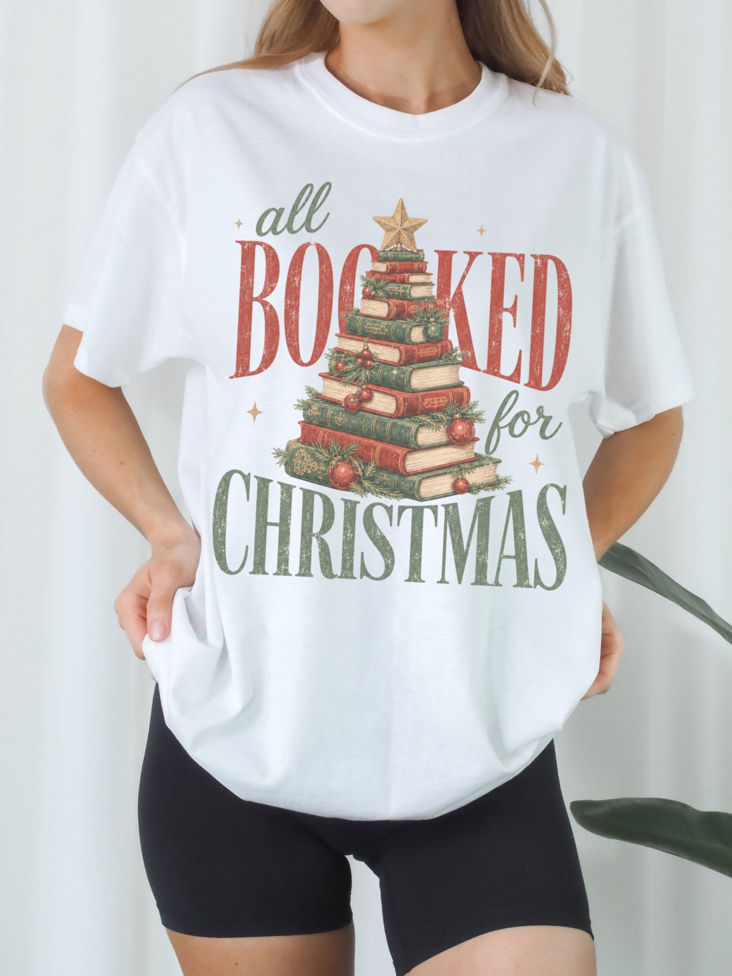 White Comfort Colors shirt modeled with an All Booked for Christmas graphic featuring a Christmas tree made of stacked red and green books with evergreen branches, ornaments and gold star, festive Christmas shirt for readers