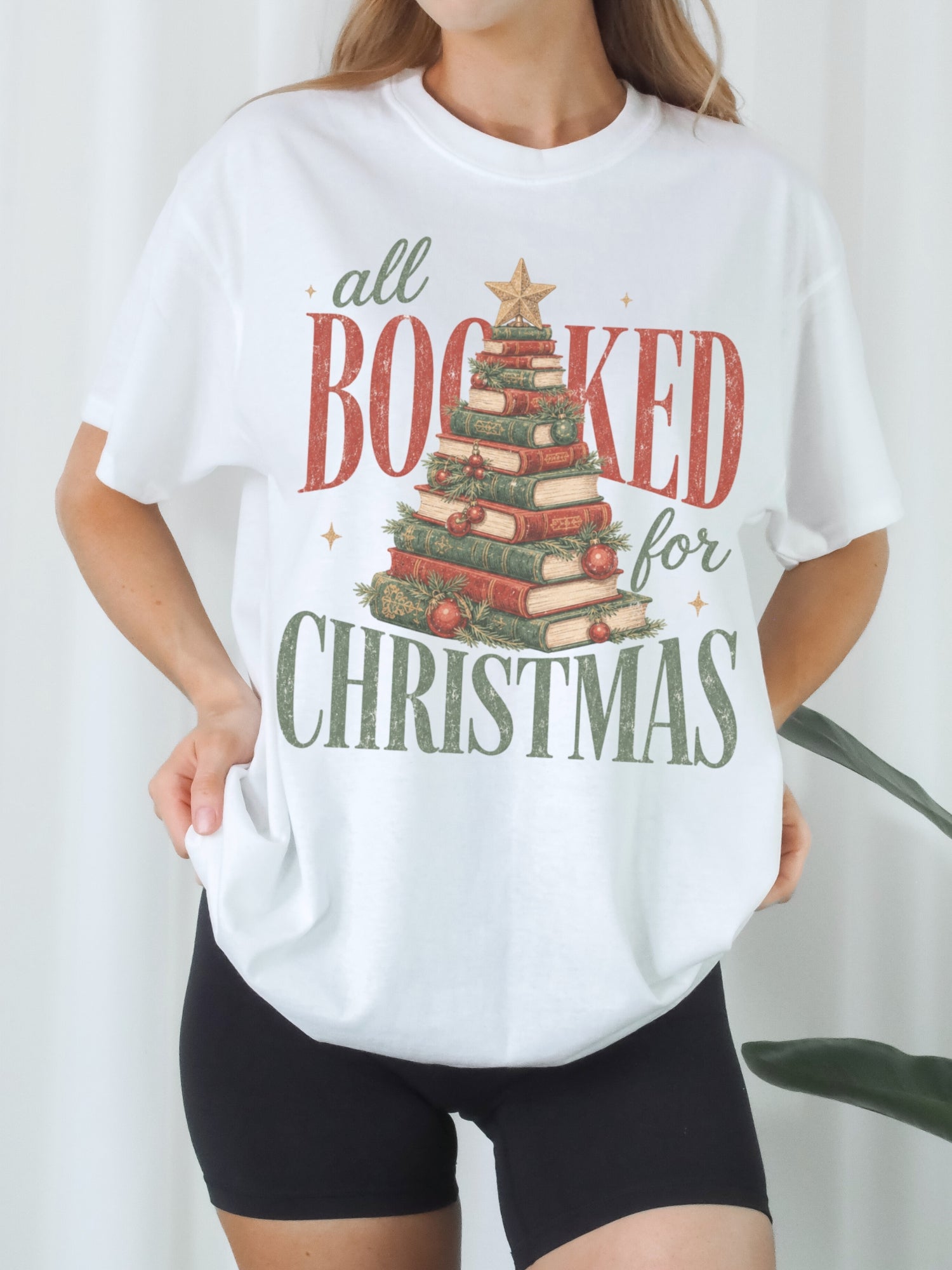 White Comfort Colors shirt modeled with an All Booked for Christmas graphic featuring a Christmas tree made of stacked red and green books with evergreen branches, ornaments and gold star, festive Christmas shirt for readers