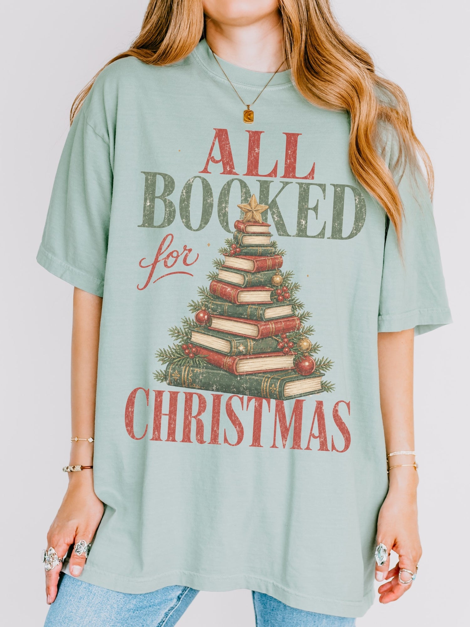 Bay Comfort Colors shirt featuring an All Booked for Christmas design with a Christmas tree made from stacked vintage books, evergreen branches, red and gold ornaments, berries and a gold star, festive bookish Christmas shirt for readers