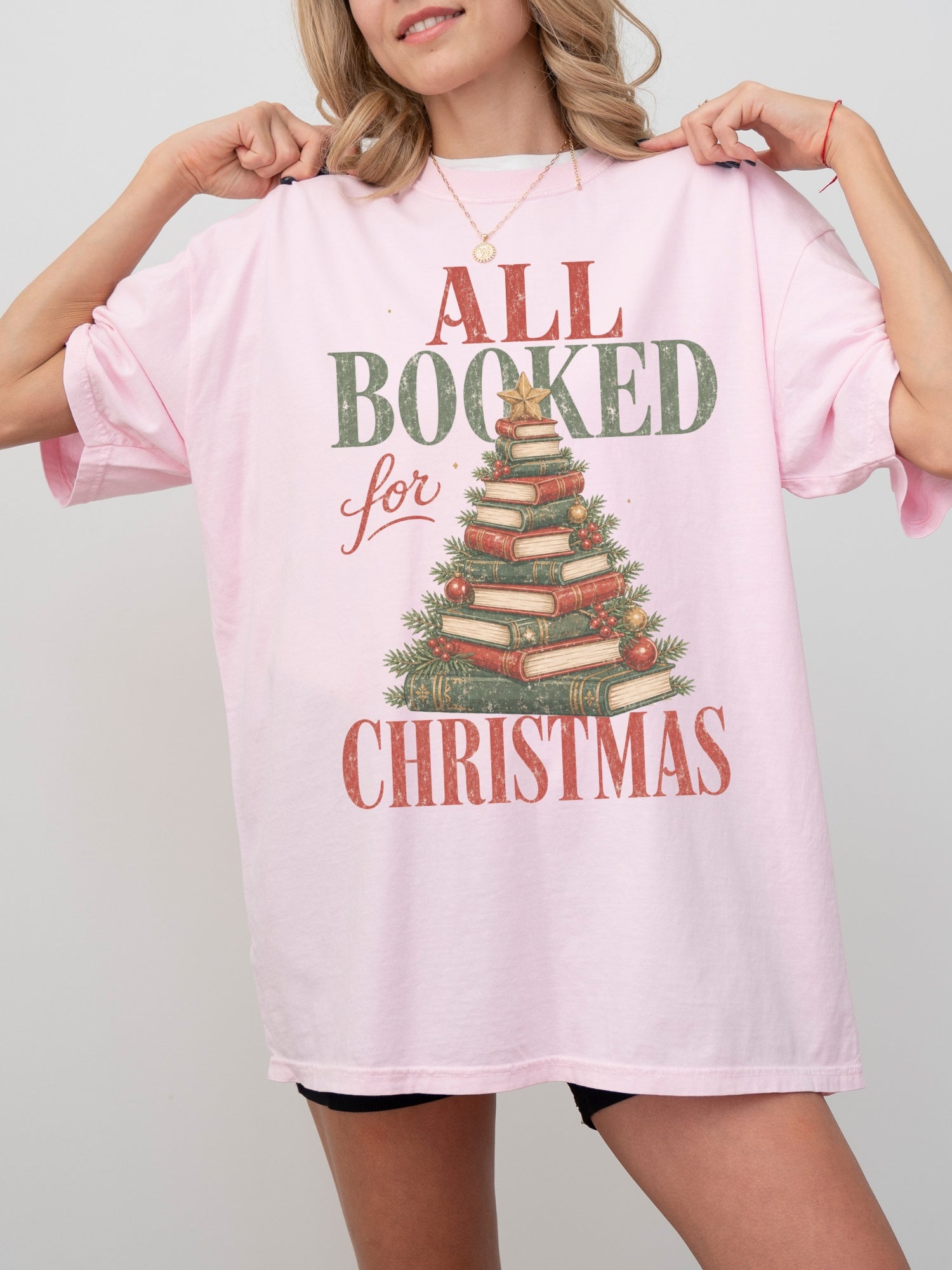 Blossom Comfort Colors shirt featuring an All Booked for Christmas graphic with stacked red and green books arranged as a Christmas tree with evergreen branches, ornaments, berries and gold star, holiday book lover shirt