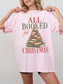 Blossom Comfort Colors shirt featuring an All Booked for Christmas graphic with stacked red and green books arranged as a Christmas tree with evergreen branches, ornaments, berries and gold star, holiday book lover shirt