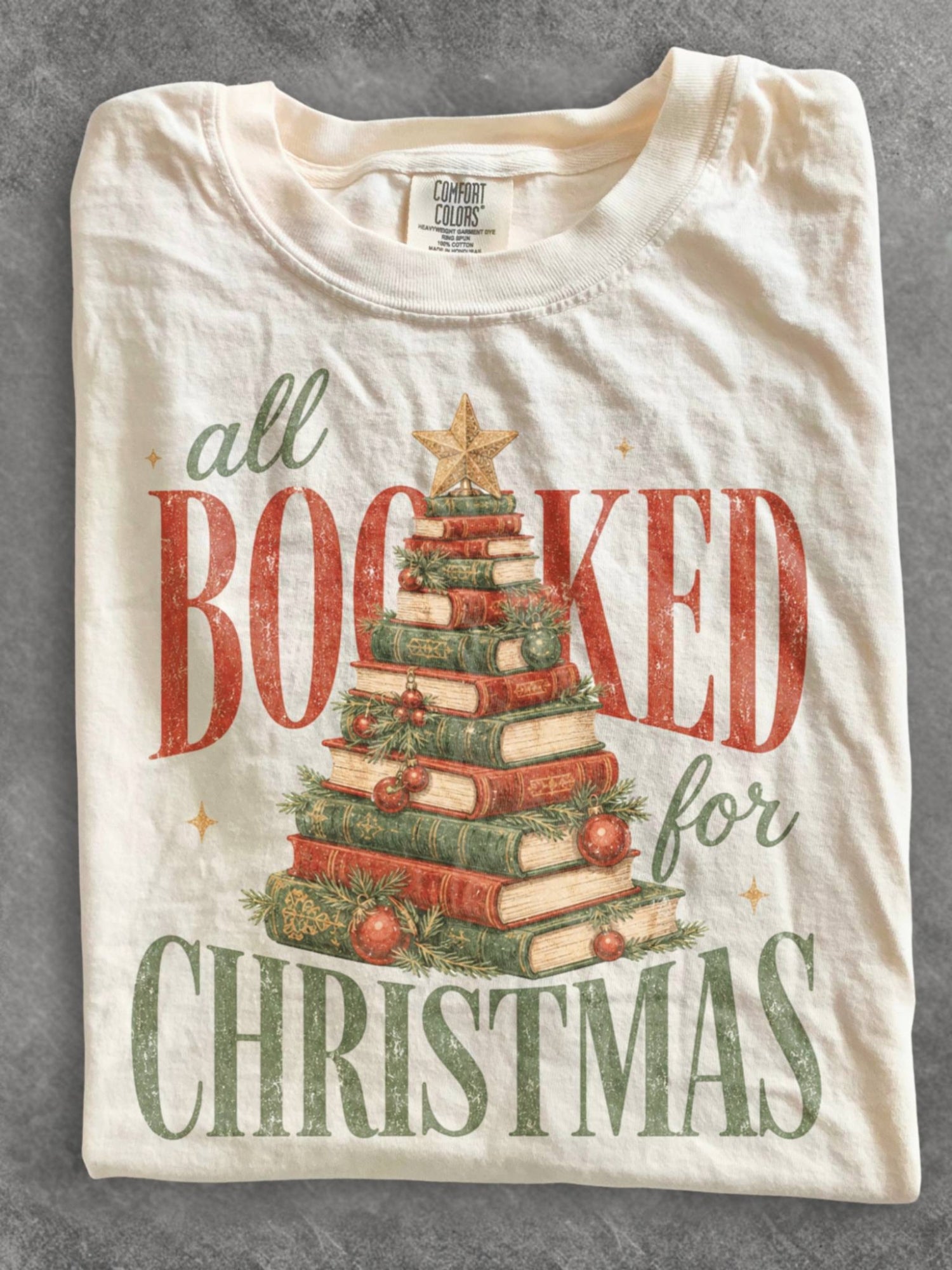Ivory Comfort Colors shirt featuring an All Booked for Christmas design with a book Christmas tree made from stacked vintage books decorated with evergreen branches, red ornaments and a gold star, festive holiday shirt for book lovers