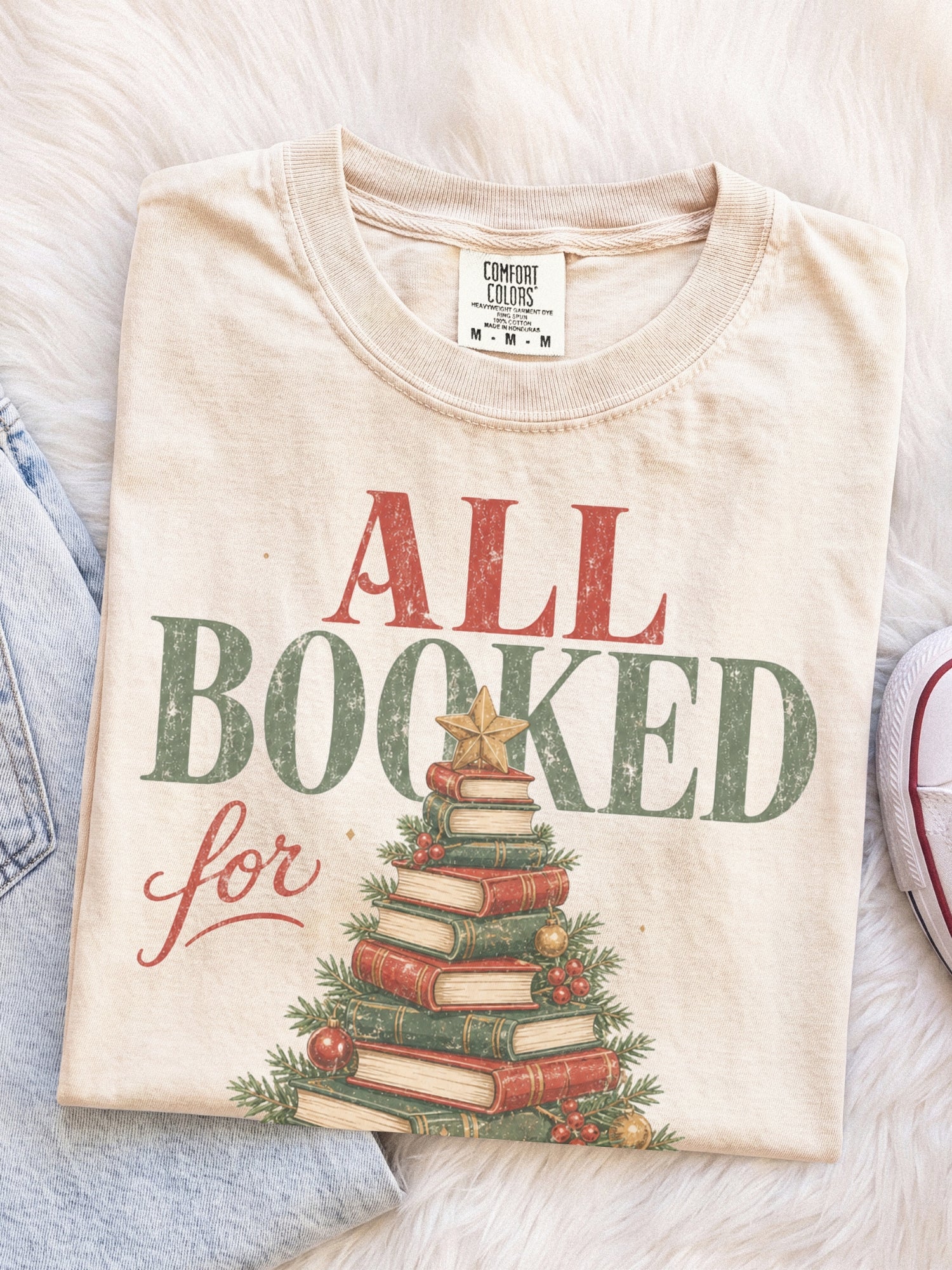 Ivory Comfort Colors shirt featuring an All Booked for Christmas graphic with a Christmas tree made from stacked vintage books decorated with evergreen branches, red berries, ornaments and a gold star, cozy Christmas reader shirt