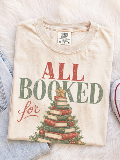 Ivory Comfort Colors shirt featuring an All Booked for Christmas graphic with a Christmas tree made from stacked vintage books decorated with evergreen branches, red berries, ornaments and a gold star, cozy Christmas reader shirt