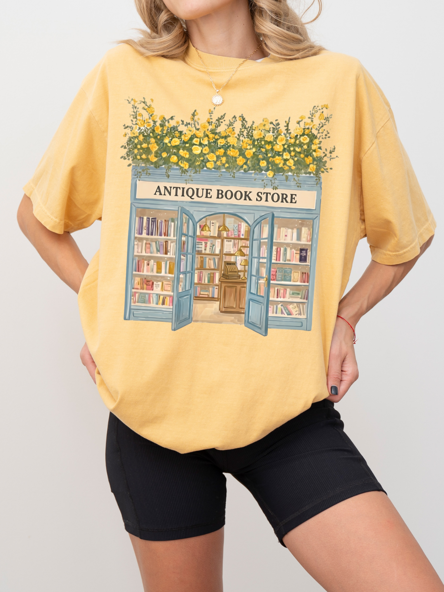 Woman wearing a mustard t shirt with a vintage bookstore illustration featuring floral greenery, open doors, and shelves of books styled in a relaxed pose