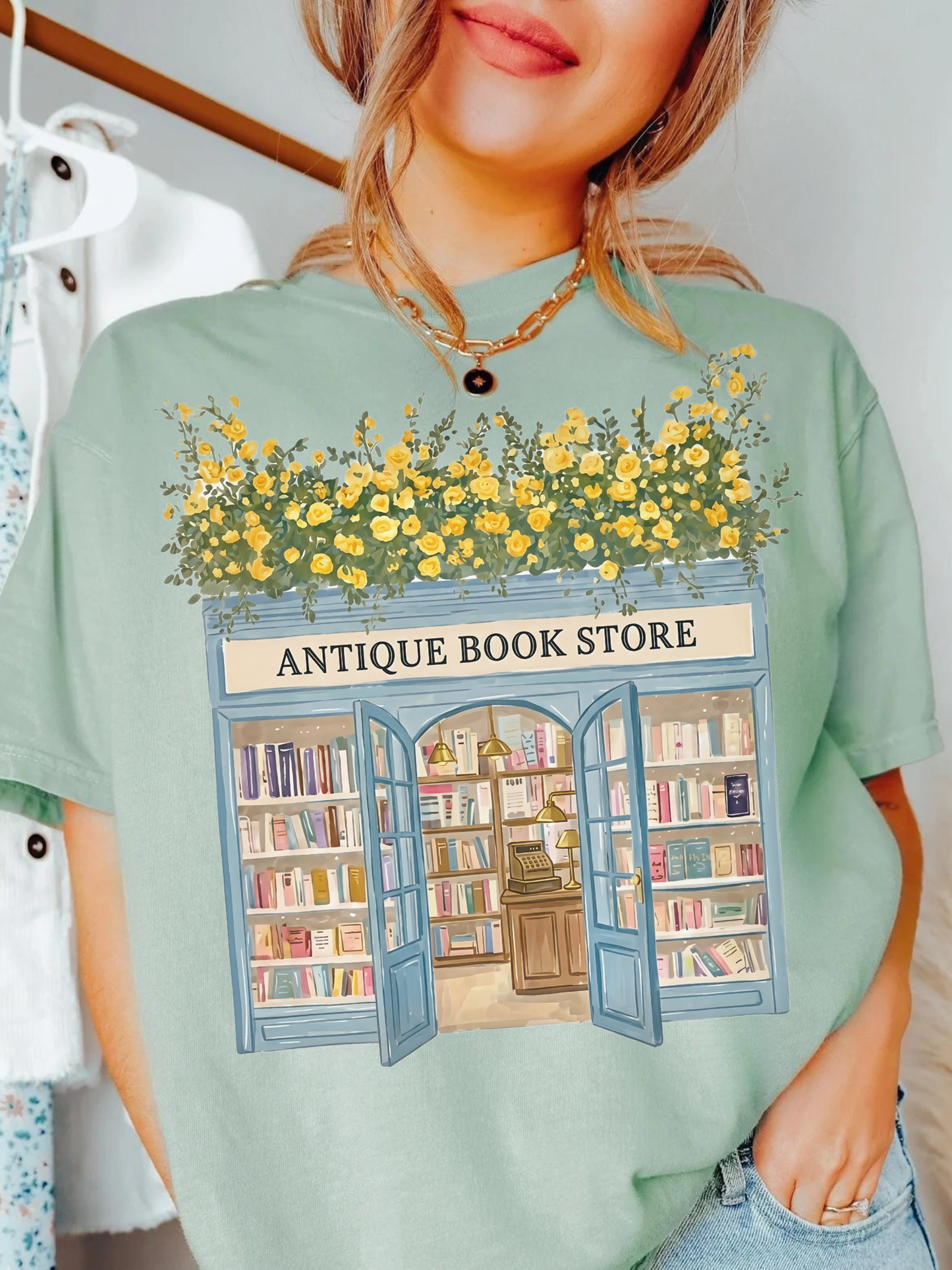Woman wearing a bay green t shirt with a vintage antique bookstore illustration featuring open doors, bookshelves, and yellow flowers on top styled with gold jewelry in a cozy indoor setting