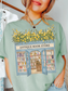 Woman wearing a bay green t shirt with a vintage antique bookstore illustration featuring open doors, bookshelves, and yellow flowers on top styled with gold jewelry in a cozy indoor setting