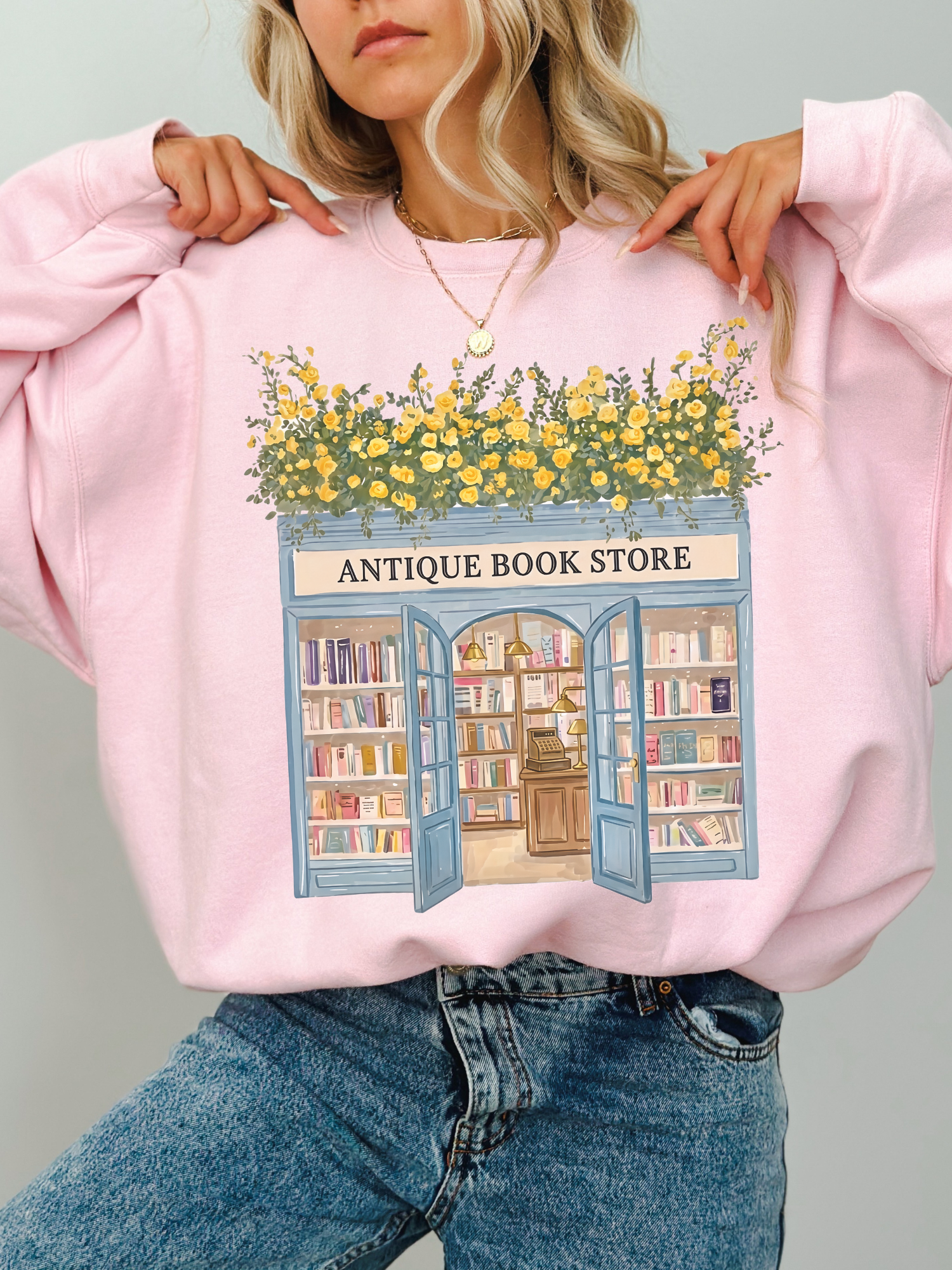 Woman wearing a pink sweatshirt with a cozy bookstore graphic featuring open doors, stacked books, and yellow flowers styled with denim jeans and layered jewelry