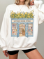 Woman wearing a white colored sweatshirt with a vintage bookstore illustration featuring open doors, stacked books, and yellow floral greenery styled in a cozy casual outfit