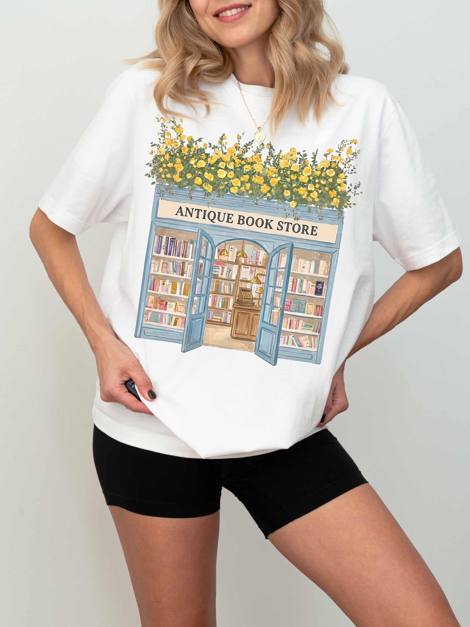 Woman wearing a white t shirt with an antique bookstore illustration featuring open doors, bookshelves, and yellow flowers styled in a clean minimal setting