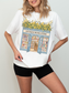Woman wearing a white t shirt with an antique bookstore illustration featuring open doors, bookshelves, and yellow flowers styled in a clean minimal setting