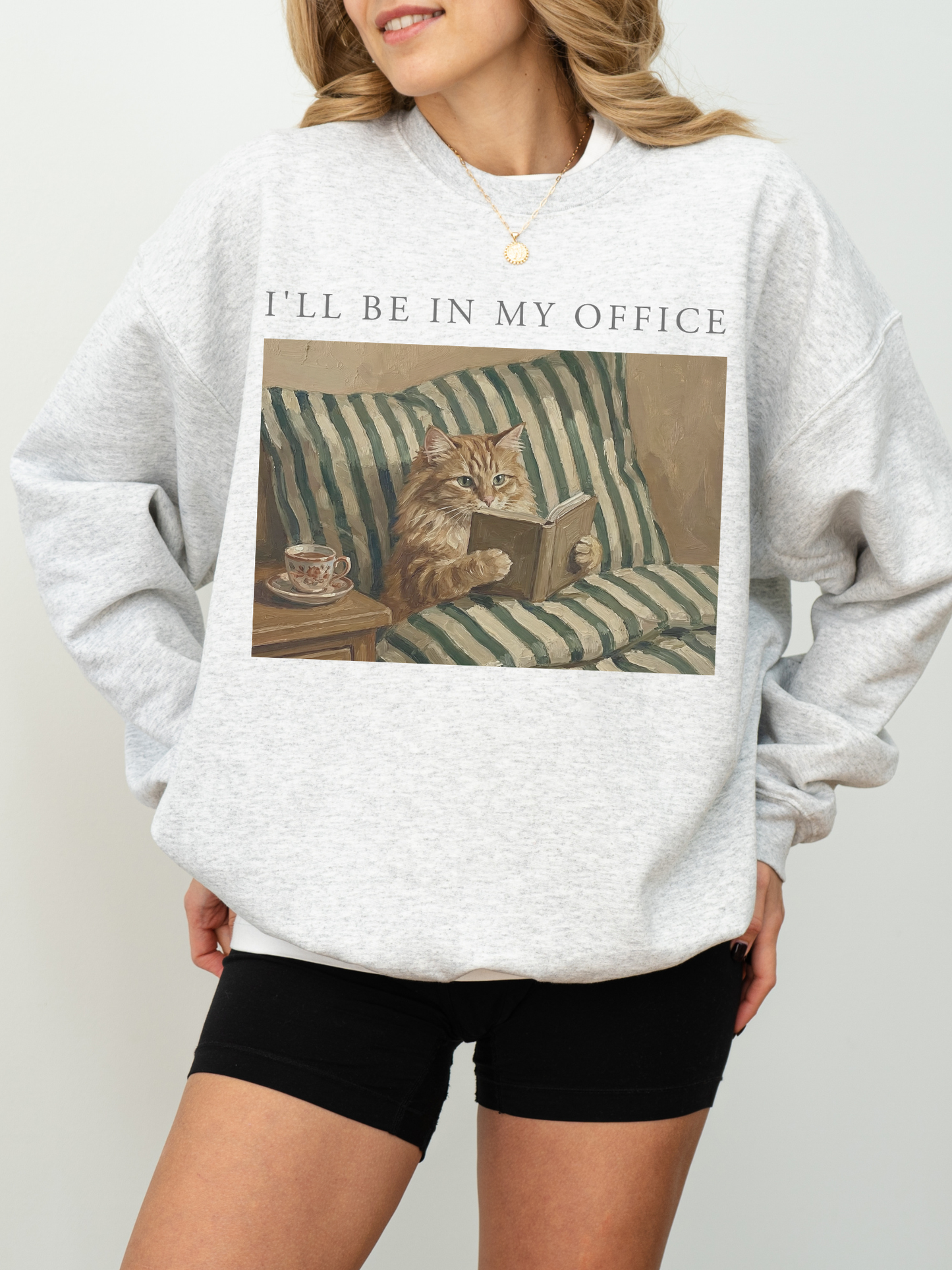 Light grey oversized sweatshirt featuring Ill be in my office text and a vintage painting of a cat reading a book in bed beside a tea cup. Relaxed cozy outfit for readers, book lovers, and cat themed apparel fans.