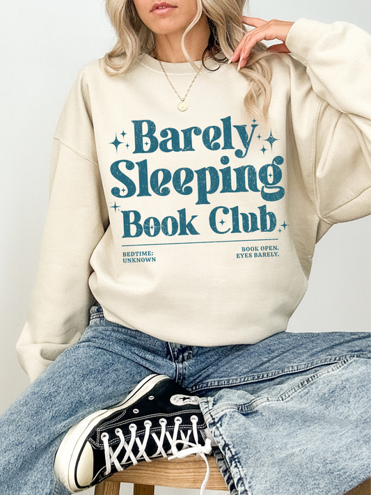 Woman wearing a sand Barely Sleeping Book Club sweatshirt with blue vintage reading design