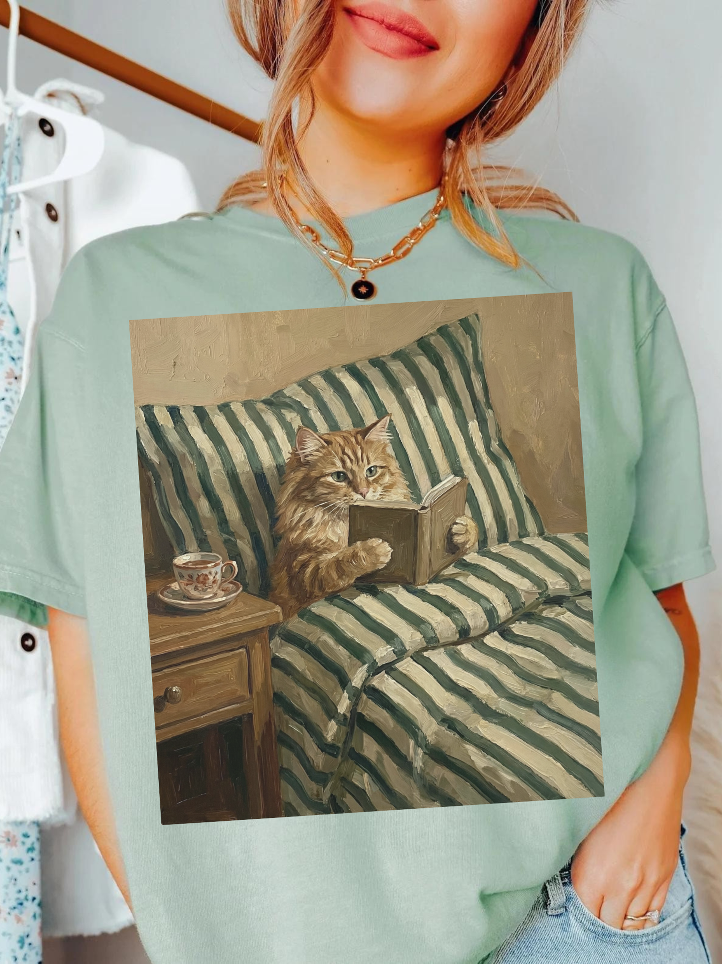 Sage green oversized t-shirt with a vintage painting style cat reading a book in bed beside a tea cup on a nightstand. Styled on a model indoors with a soft cozy aesthetic, perfect for readers, book lovers, and cat themed fashion.