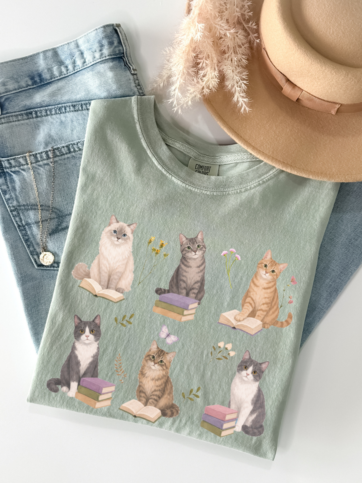 Flat lay of bay green Comfort Colors t shirt featuring illustrated cats reading books with floral accents paired with denim jeans necklace and neutral hat