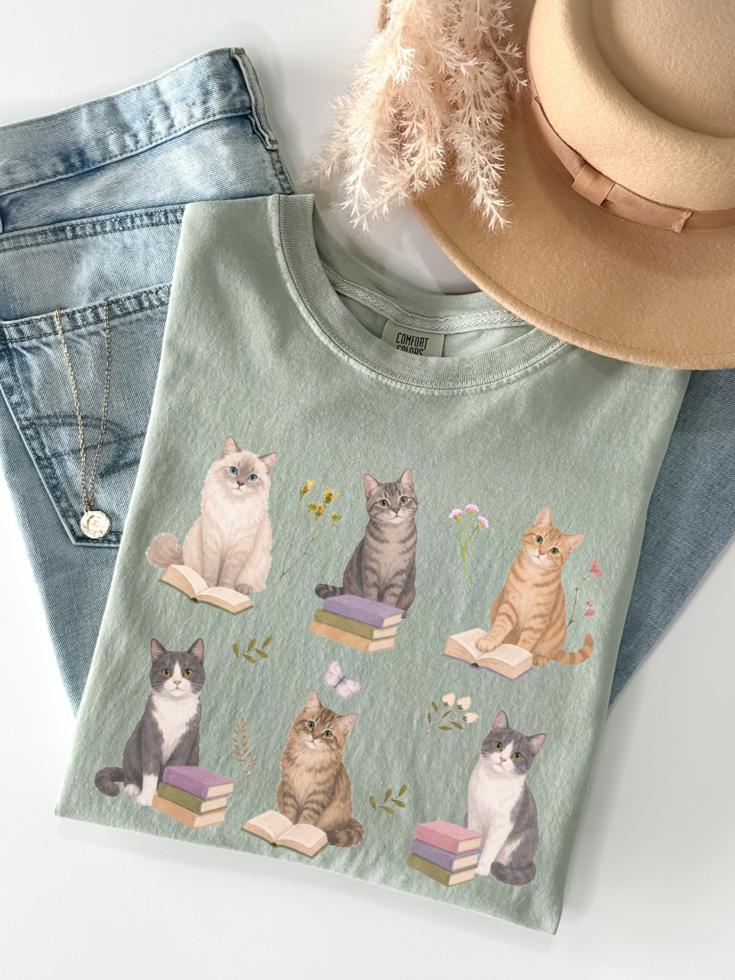 Flat lay of bay green Comfort Colors t shirt featuring illustrated cats reading books with floral accents paired with denim jeans necklace and neutral hat