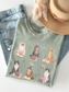 Flat lay of bay green Comfort Colors t shirt featuring illustrated cats reading books with floral accents paired with denim jeans necklace and neutral hat