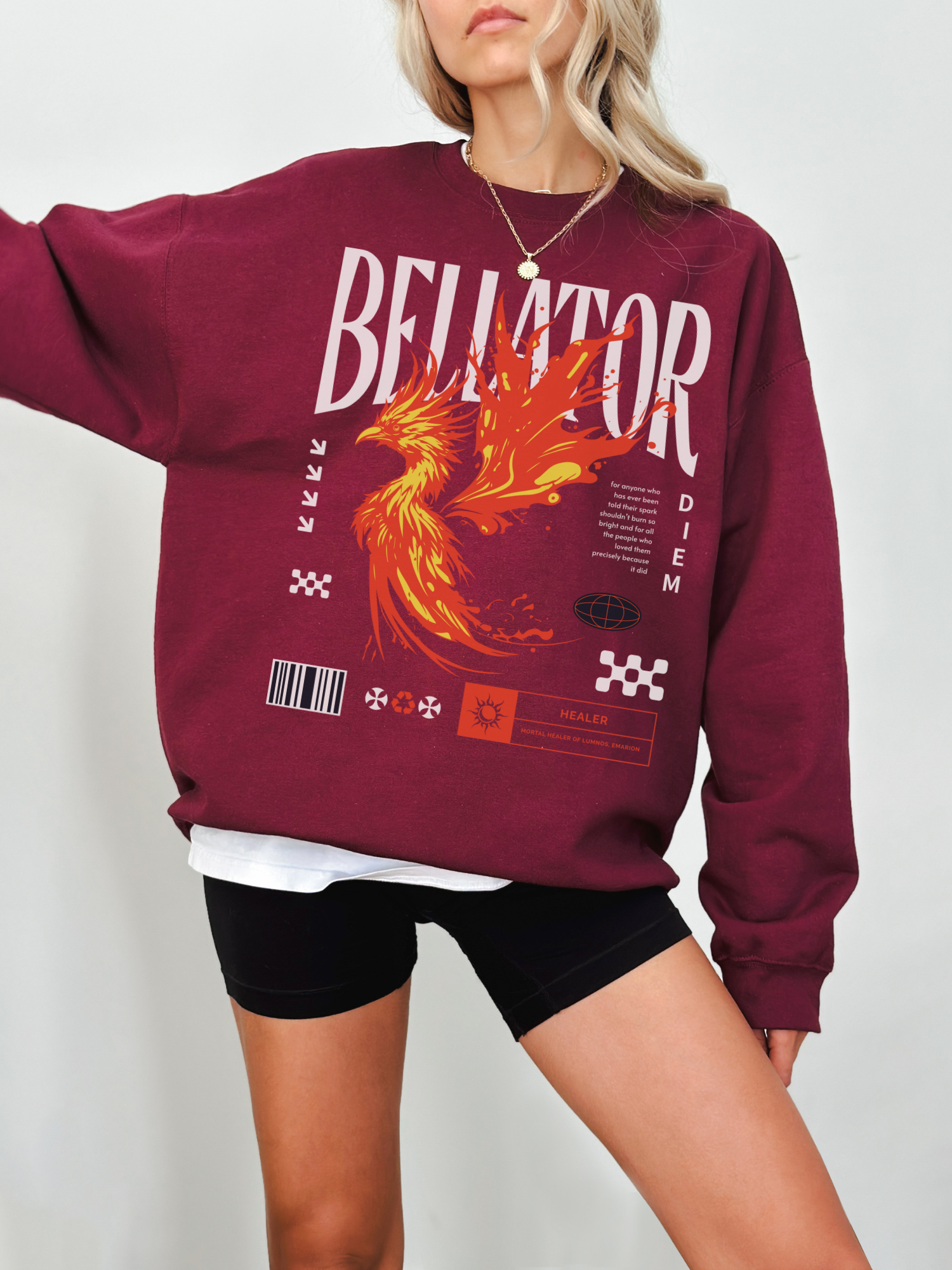 Maroon crewneck sweatshirt featuring bold phoenix fire design and Bellator fantasy text. Model standing with arm extended in front of light background, wearing layered tee and necklace.

