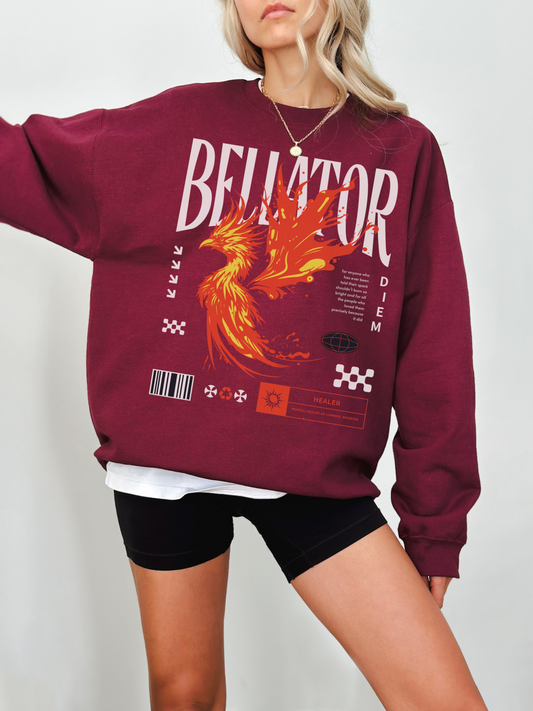 Maroon crewneck sweatshirt featuring bold phoenix fire design and Bellator fantasy text. Model standing with arm extended in front of light background, wearing layered tee and necklace.