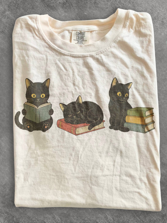 ivory comfort colors t shirt flat lay featuring three black cats with books including a reading cat sleeping cat on a book and cat beside stacked books for book lovers and cat lovers