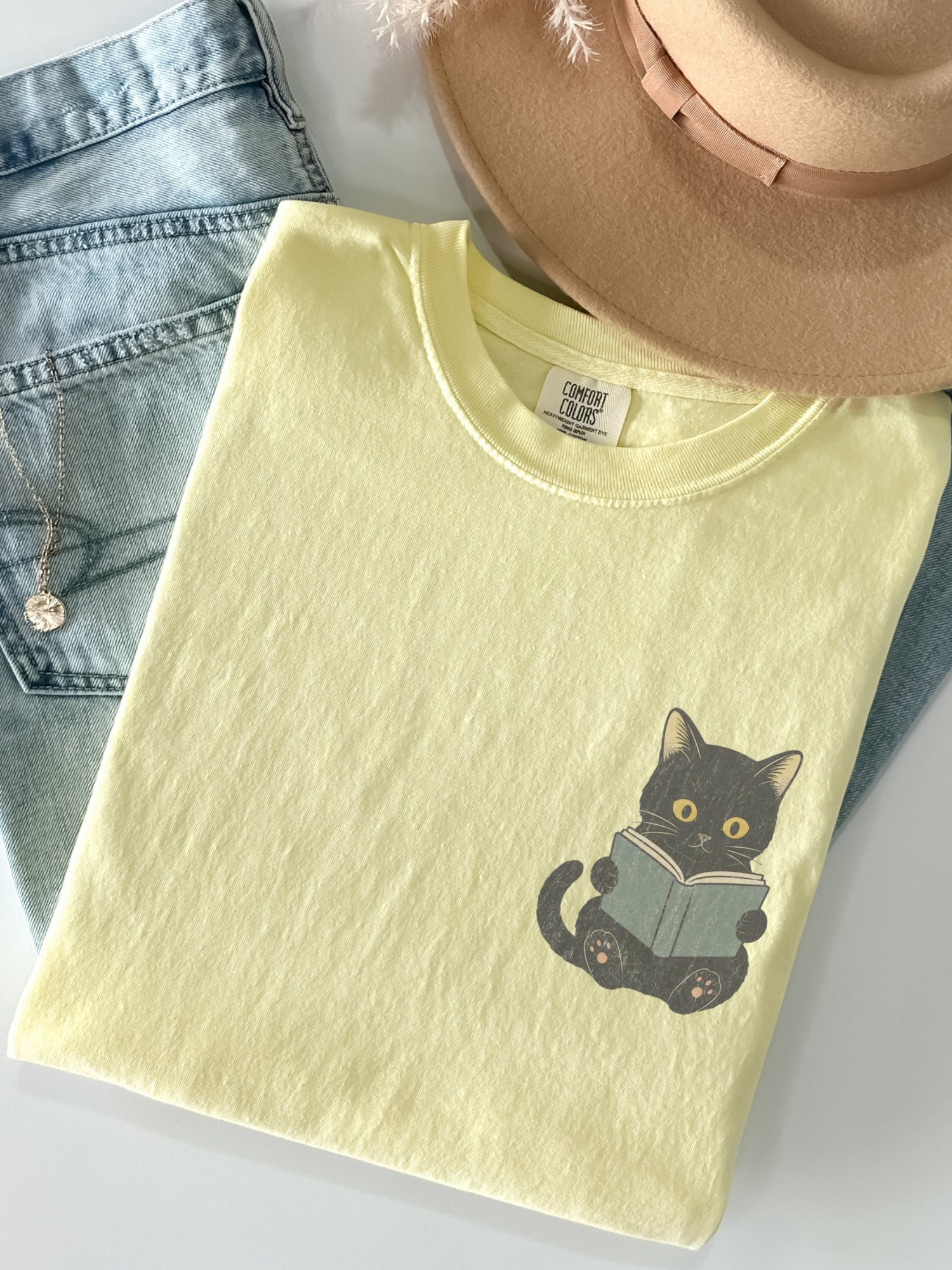 folded butter Comfort Colors shirt featuring a black cat holding a book graphic, aesthetic reading shirt for book lovers, teachers, and cat enthusiasts
